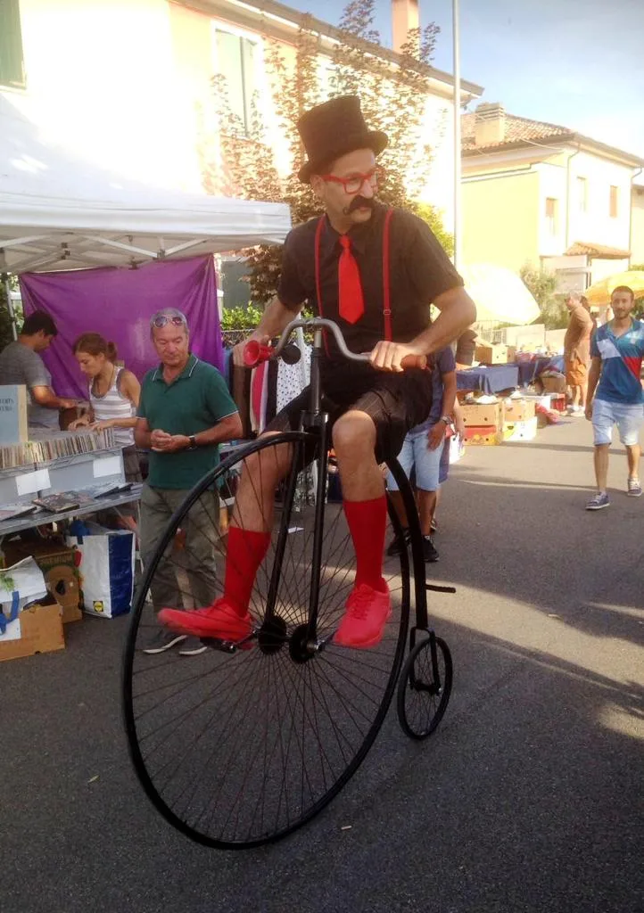 Ernesto in bombetta e cravatta rossa in sella al velocipede dalla ruota grande, in mezzo alle bancarelle