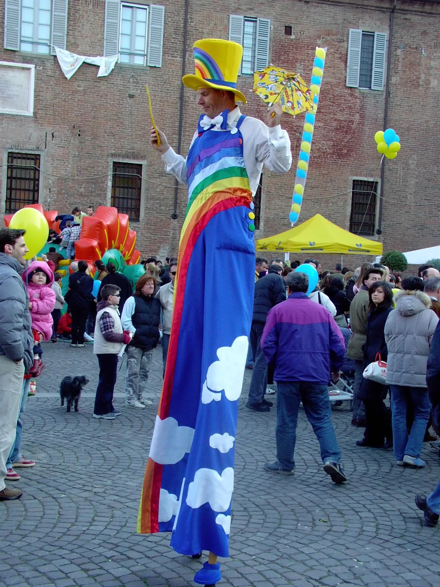 Trampoliere vestito da arcobaleno con cappello giallo che cammina sui trampoli in mezzo alla gente in piazza