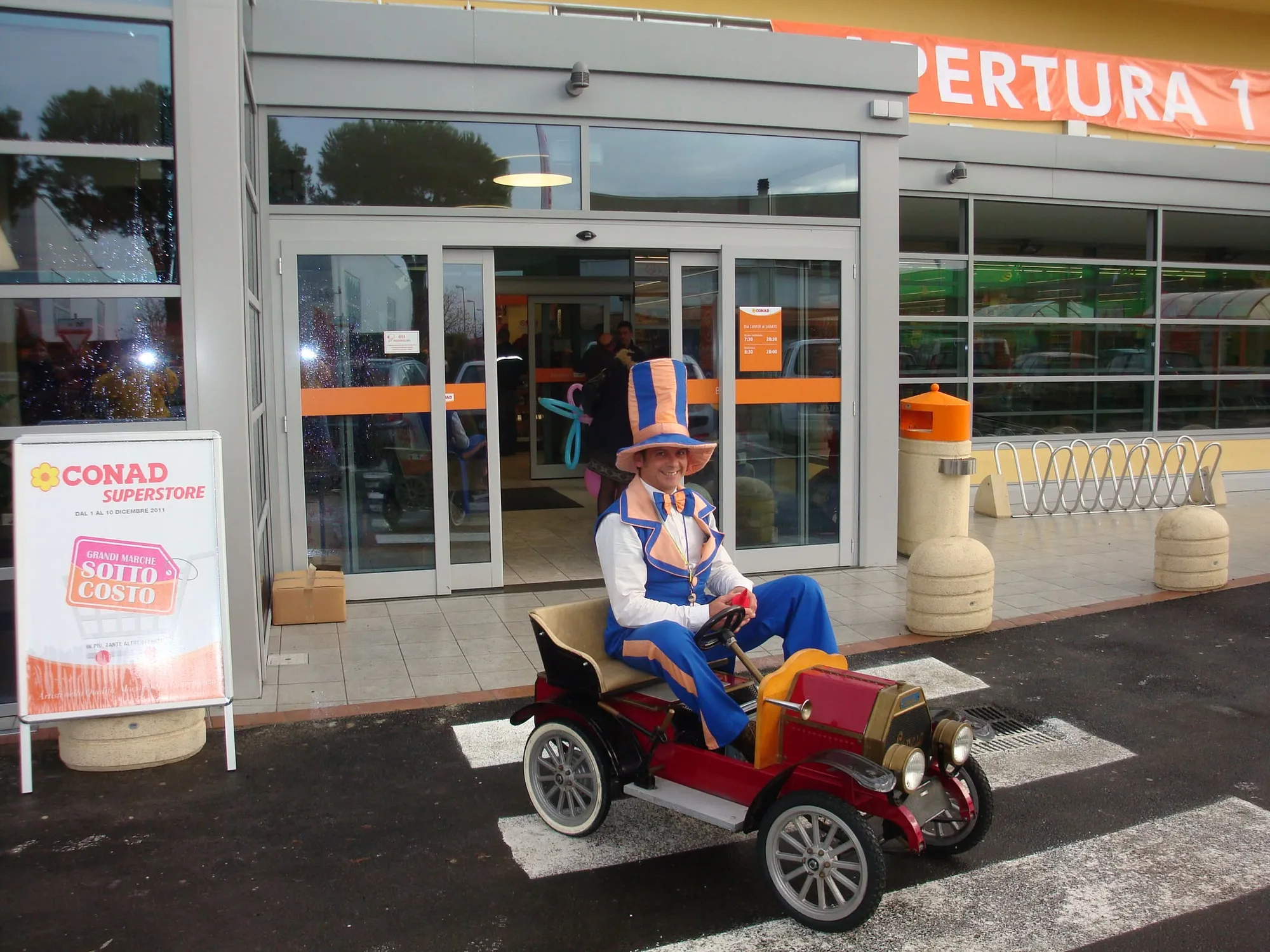 Chicco con cilindro e completo a righe blu e arancio seduto sulla sua automobilina d'epoca rossa, davanti all'ingresso di un supermercato