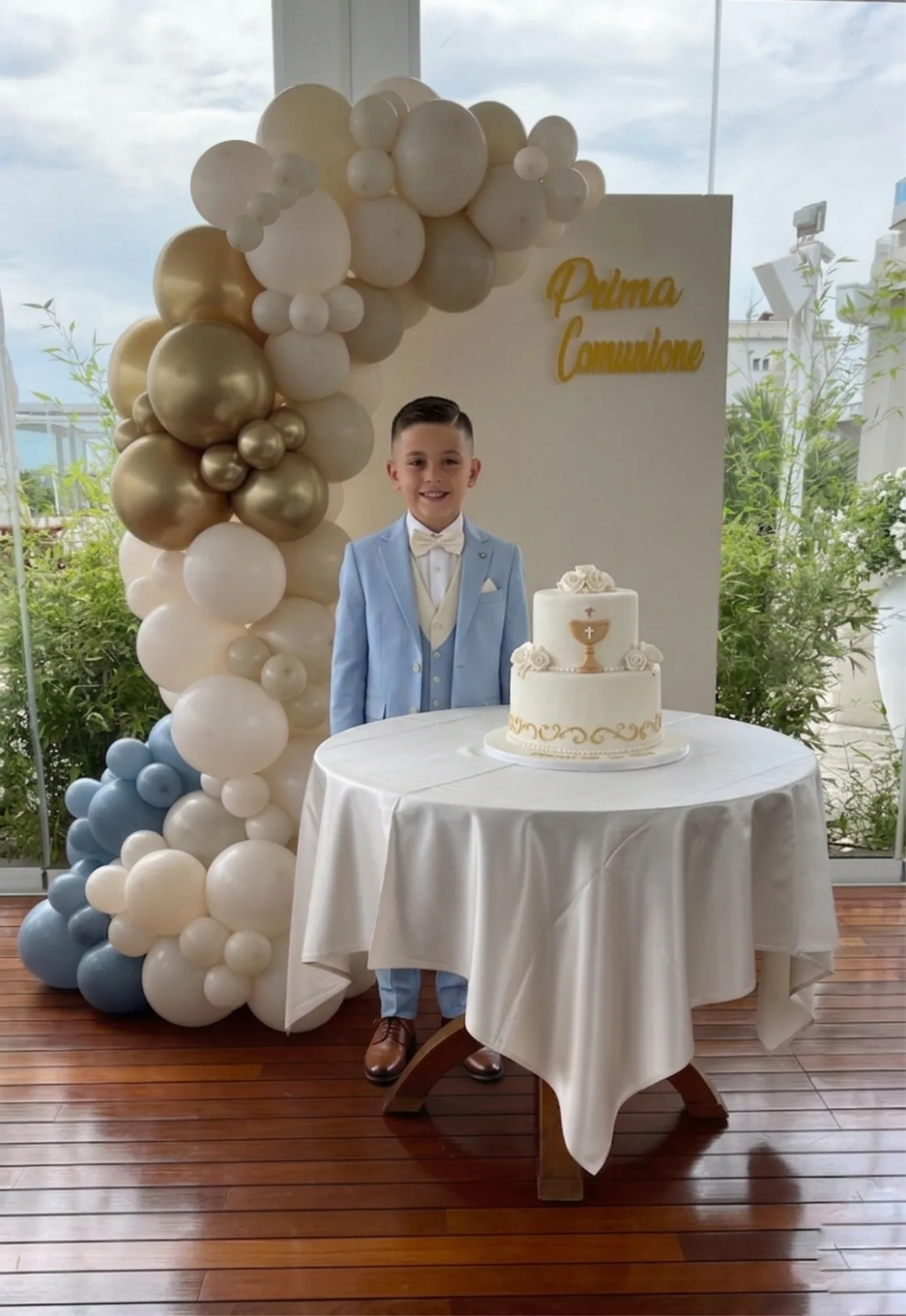 Bambino in abito celeste davanti a un arco di palloncini panna, oro e azzurro con il pannello Prima Comunione e la torta a due piani sul tavolino