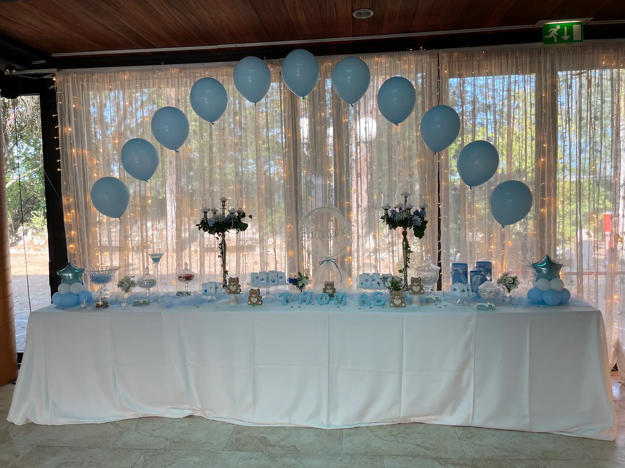 Tavolo del battesimo di Thomas con arco di palloncini azzurri sopra la tenda luminosa, confettata, lettere col nome e cerchio con la scritta Battesimo