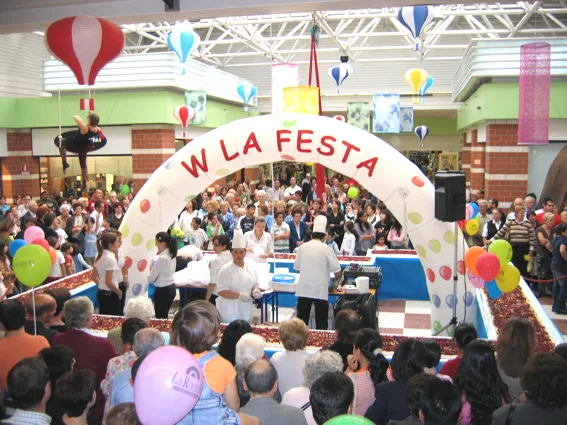 Arco gonfiabile bianco con la scritta «W la festa» sopra un palco affollato in una galleria commerciale