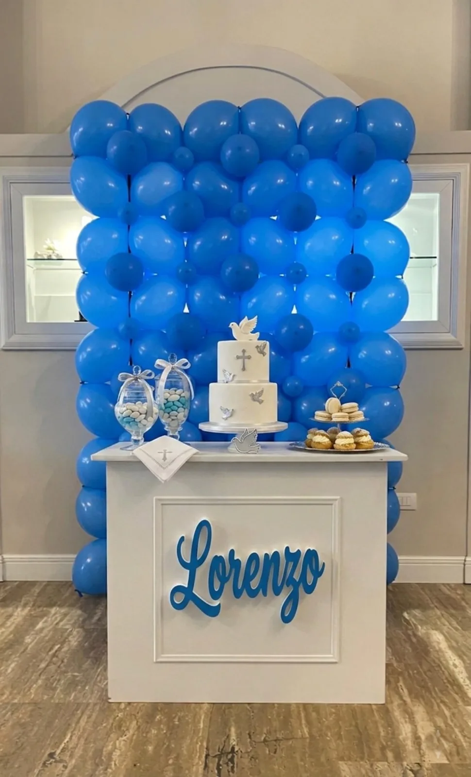 Parete di palloncini blu dietro al tavolo dei dolci, con il nome Lorenzo luminoso sul fronte, la torta bianca con croce e colomba e i vasi dei confetti