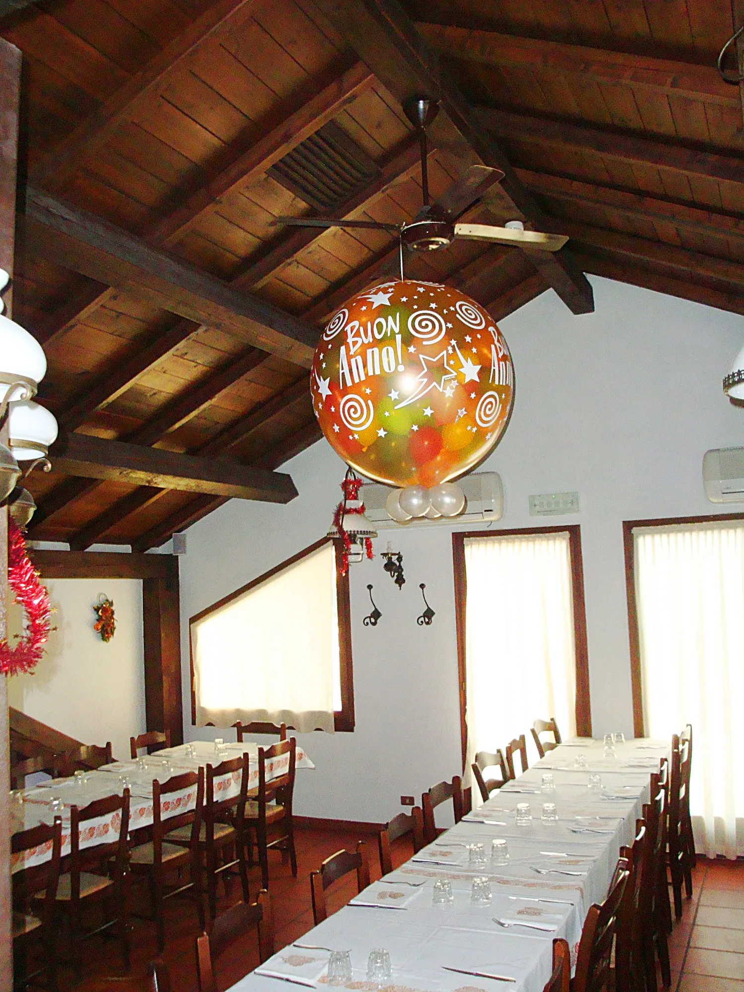 Grande palloncino dorato con la scritta Buon Anno, pieno di palloncini colorati più piccoli, appeso sopra le tavolate di una sala con il soffitto in legno