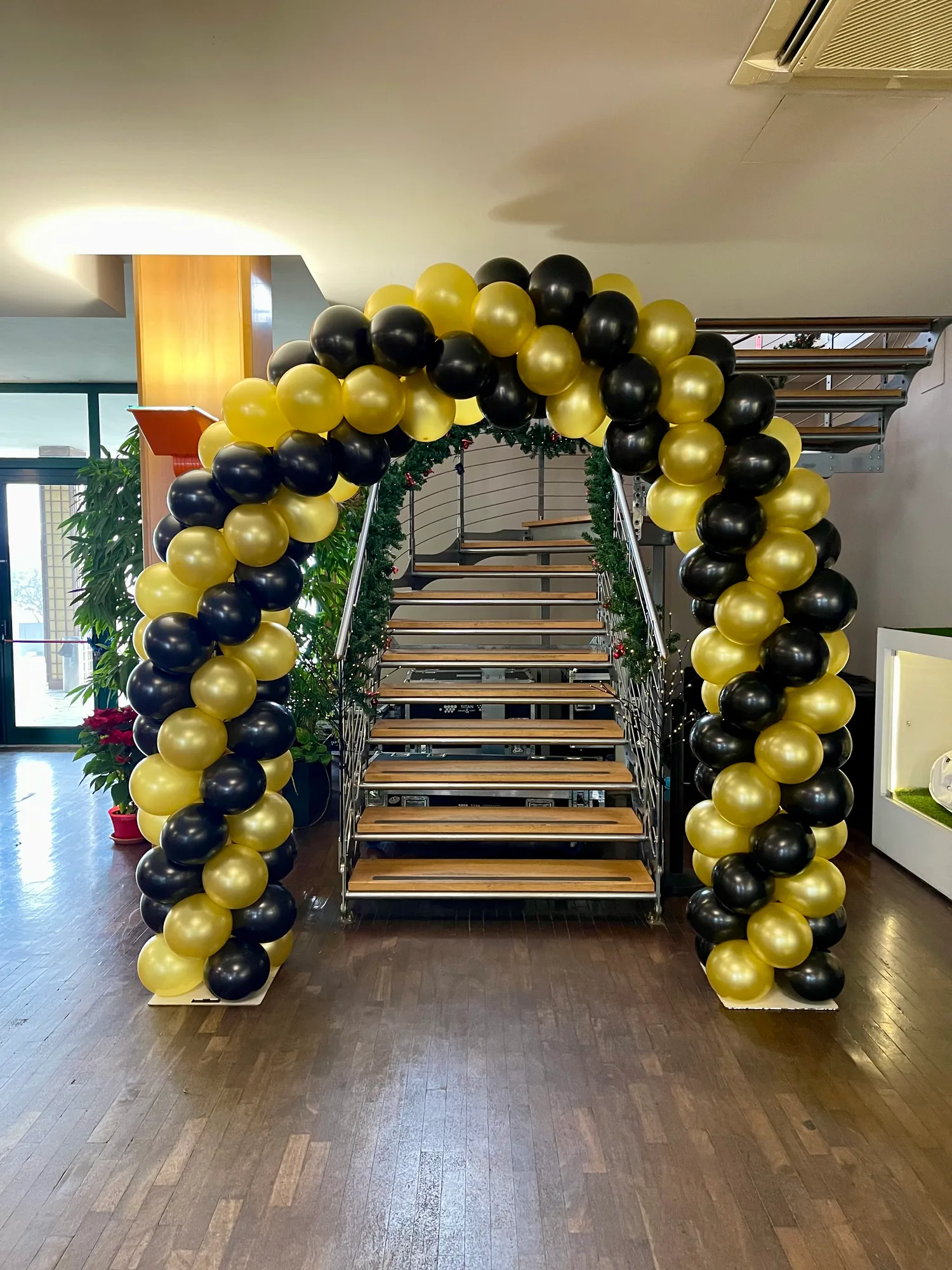 Arco di palloncini neri e oro davanti a una scala, in un locale addobbato con rami di abete