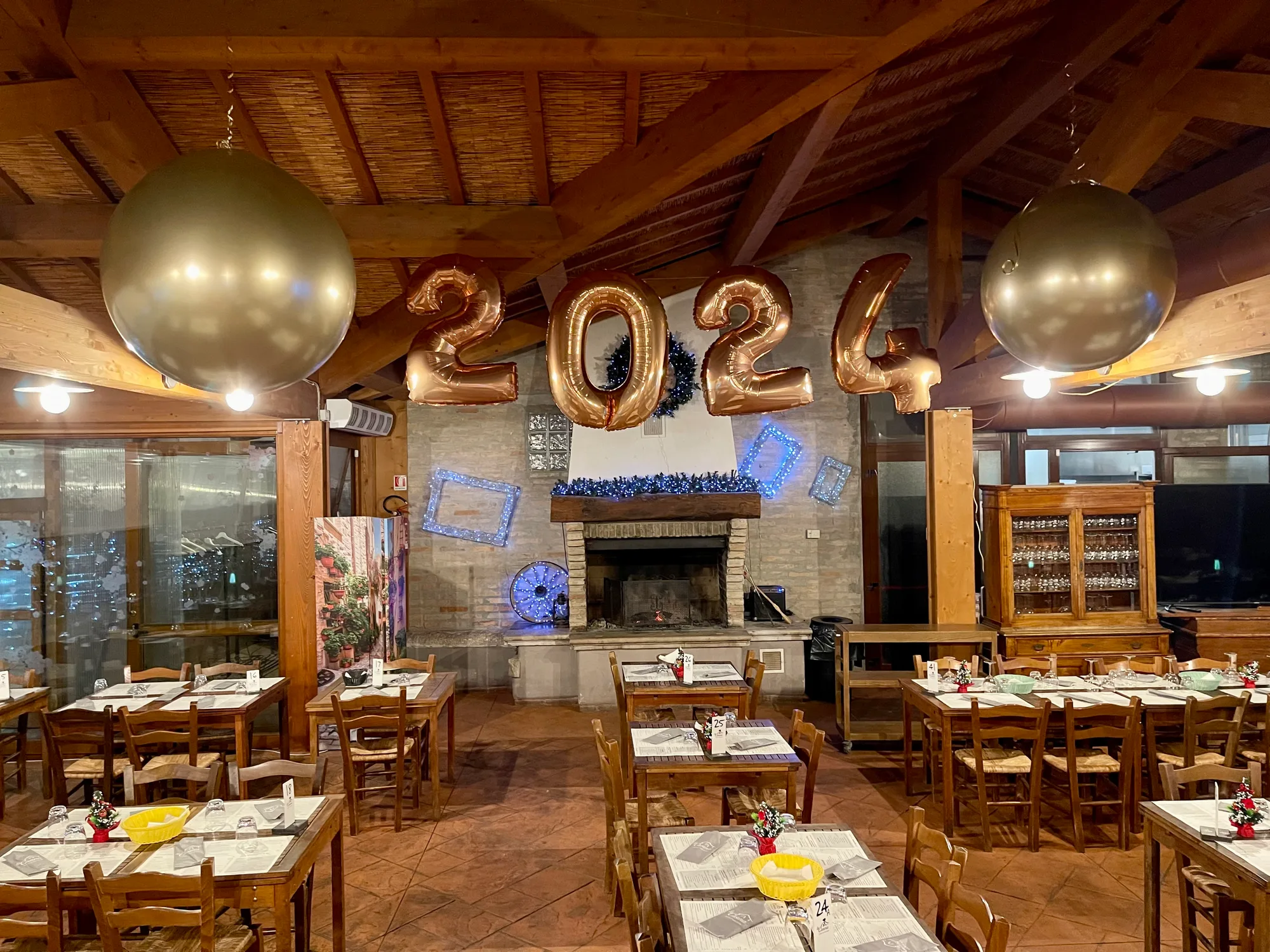 Sala di un ristorante con travi in legno addobbata per Capodanno: grandi numeri 2024 in mylar color oro rosa sopra il camino e due sfere giganti dorate appese al soffitto