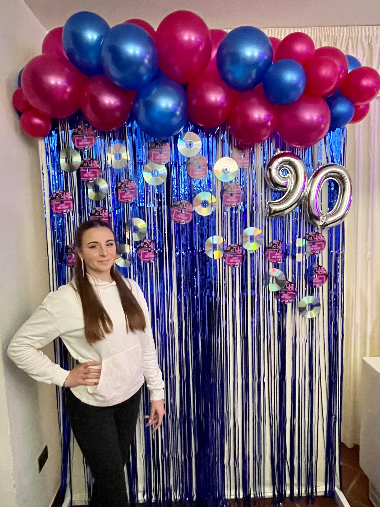 Ragazza in posa davanti a un fondale di frange blu con CD appesi, ghirlanda di palloncini fucsia e blu e numero 90 argento