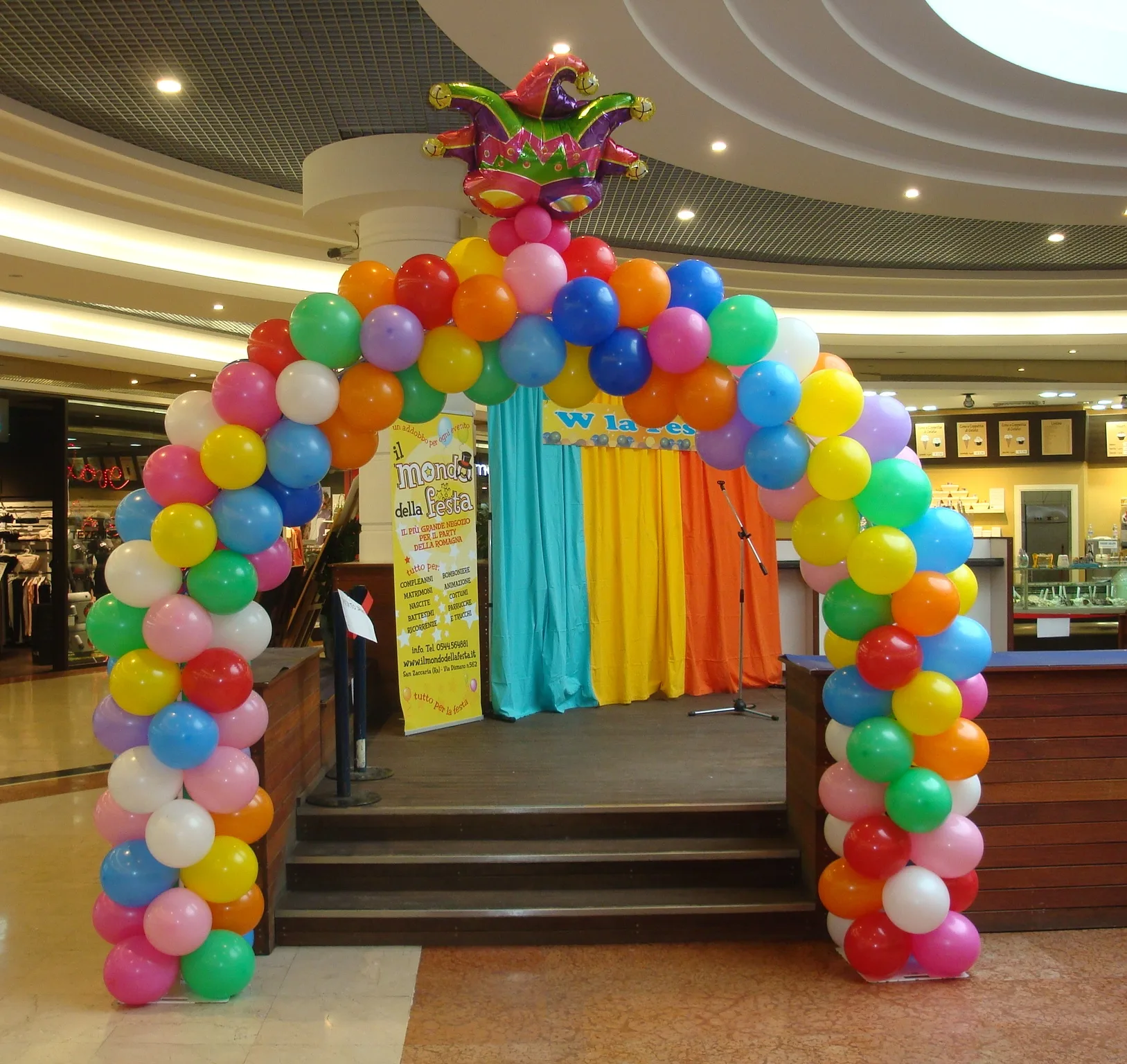 Arco di palloncini multicolore con una maschera da giullare in mylar in cima, davanti a un piccolo palco con tende colorate in una galleria commerciale