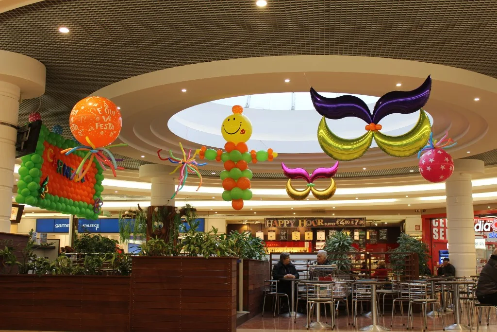 Decorazioni di Carnevale appese sopra l'area ristoro di una galleria: maschere giganti in mylar, un pagliaccio di palloncini verde e arancio, un pannello di palloncini e palloncini grandi con i nastri