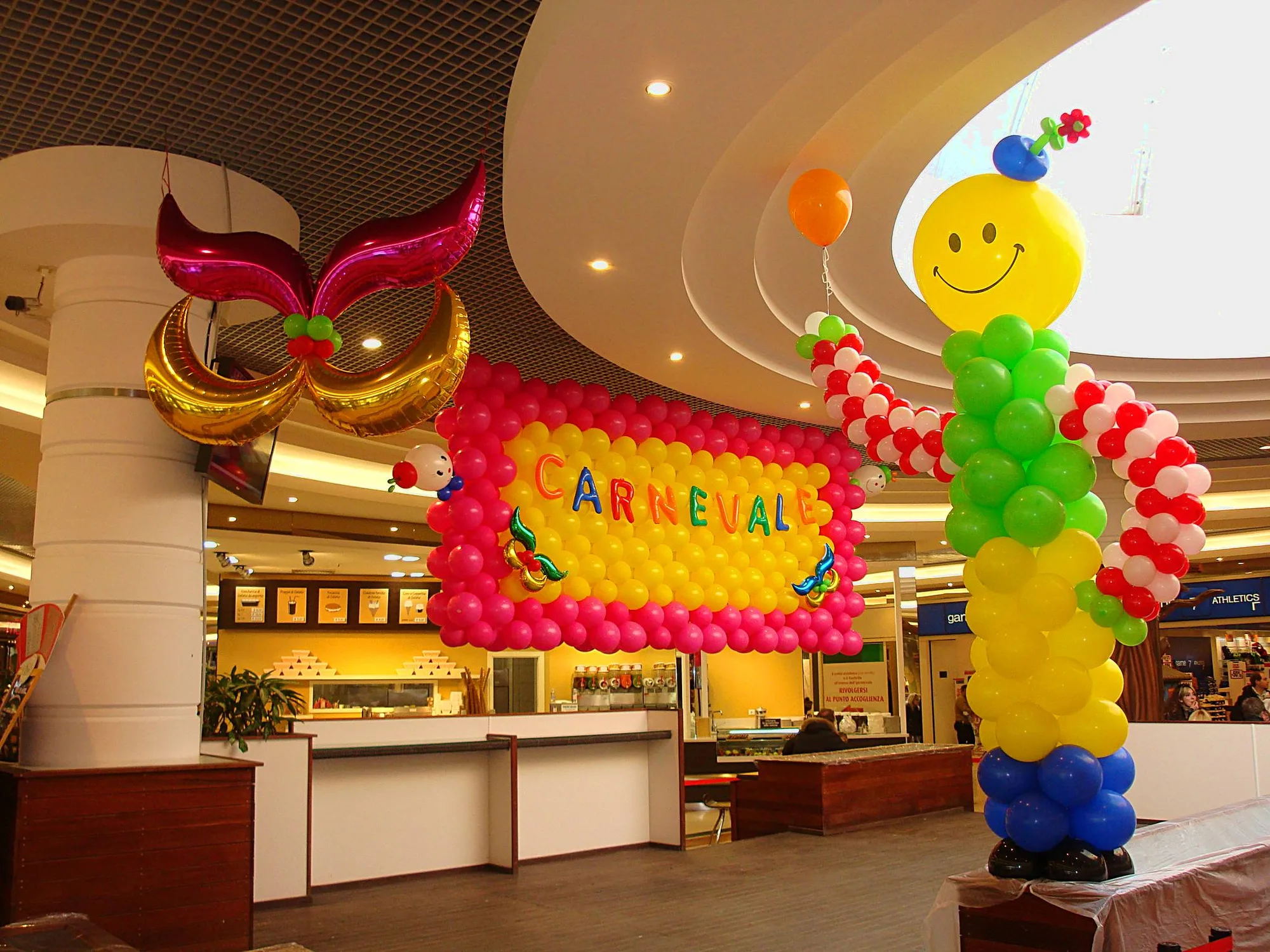 Allestimento di Carnevale in una galleria commerciale: parete di palloncini fucsia e gialli con la scritta CARNEVALE, maschera gigante in mylar fucsia e oro e un pagliaccio di palloncini con la faccina sorridente