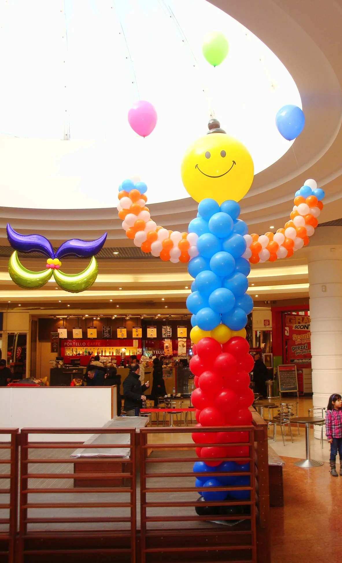 Pagliaccio di palloncini alto alcuni metri, con la testa gialla sorridente e il corpo azzurro, rosso e arancio, accanto a una maschera in mylar viola e verde appesa in una galleria