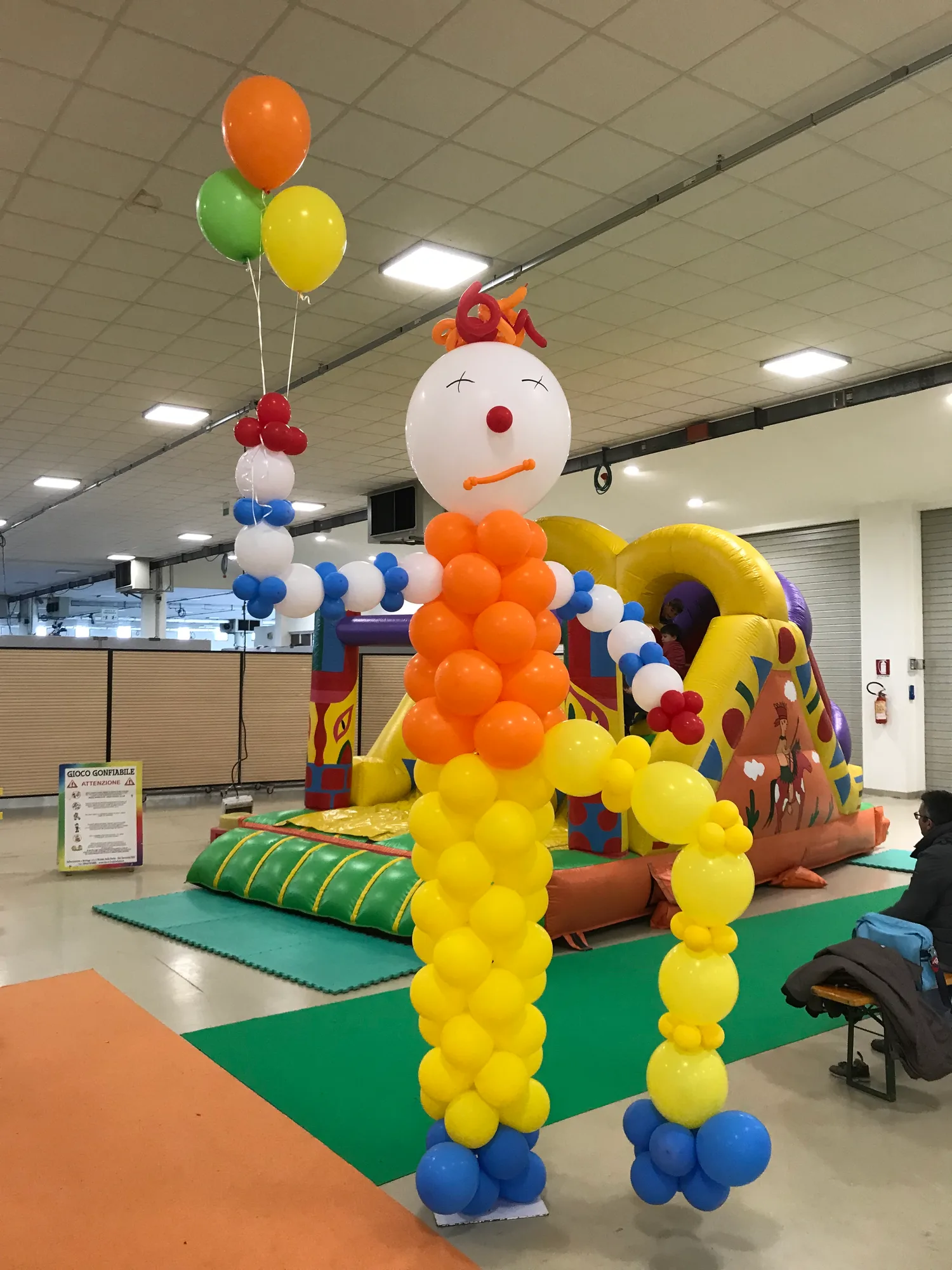 Pagliaccio di palloncini con la testa bianca, il naso rosso e un mazzo di palloncini in mano, accanto a un gioco gonfiabile in un capannone