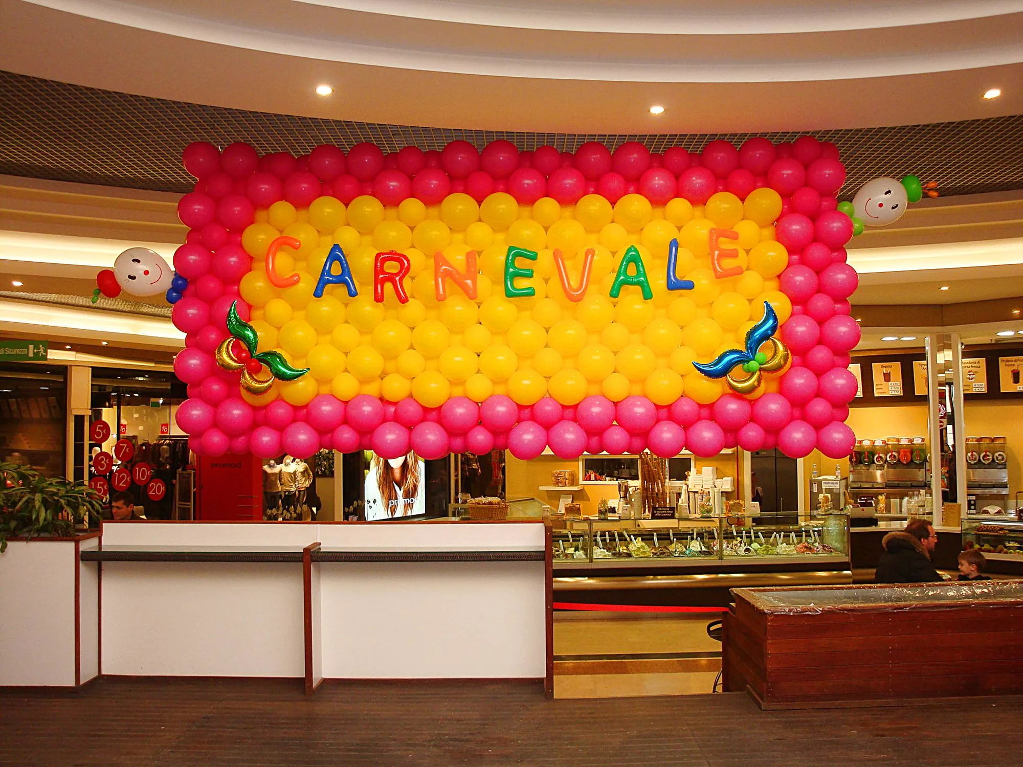 Parete di palloncini fucsia e gialli con la scritta CARNEVALE in lettere di mylar colorate, due maschere in mylar e due faccine di palloncino ai lati, sopra un bancone in galleria