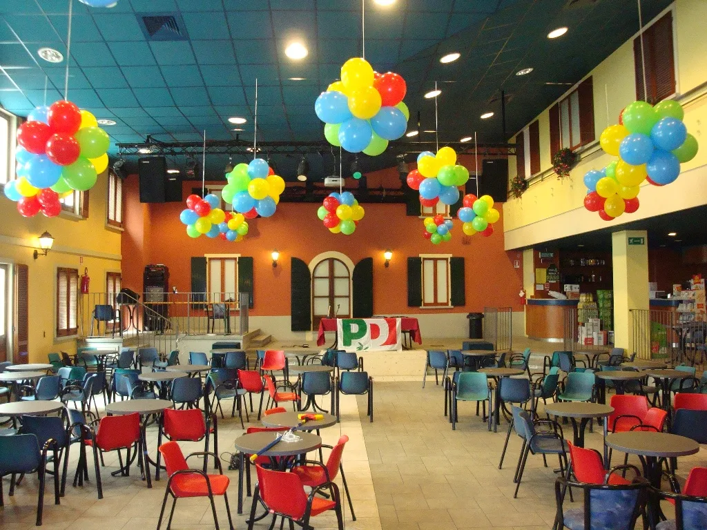 Grappoli di palloncini rossi, gialli, verdi e blu appesi al soffitto di una grande sala con tavolini e sedie colorate