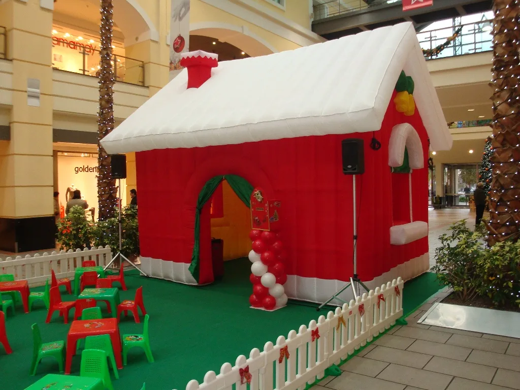 Casetta gonfiabile rossa con tetto bianco e camino, allestita in una galleria commerciale con tavolini e sedie per bambini