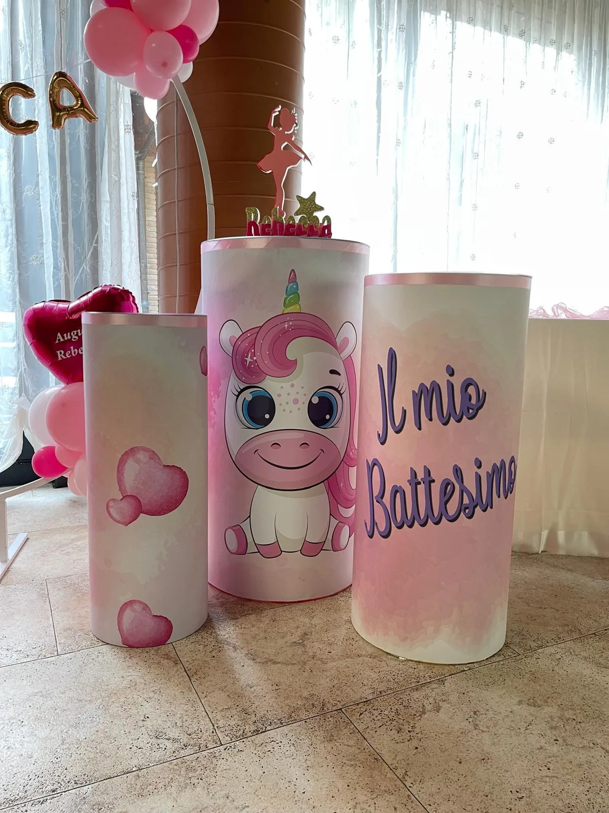 Tre cilindri bianchi e rosa con grafica di unicorno, cuori e la scritta 'Il mio Battesimo', usati come colonne per il tavolo della torta