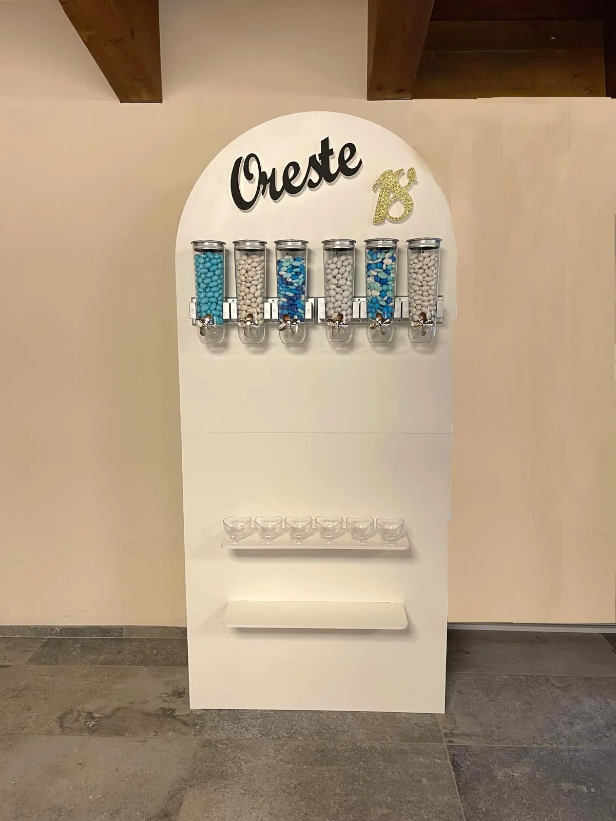 Mobile bianco ad arco con sei dispenser di confetti azzurri e bianchi, coppette a cuore e il nome Oreste con il numero 18 sopra