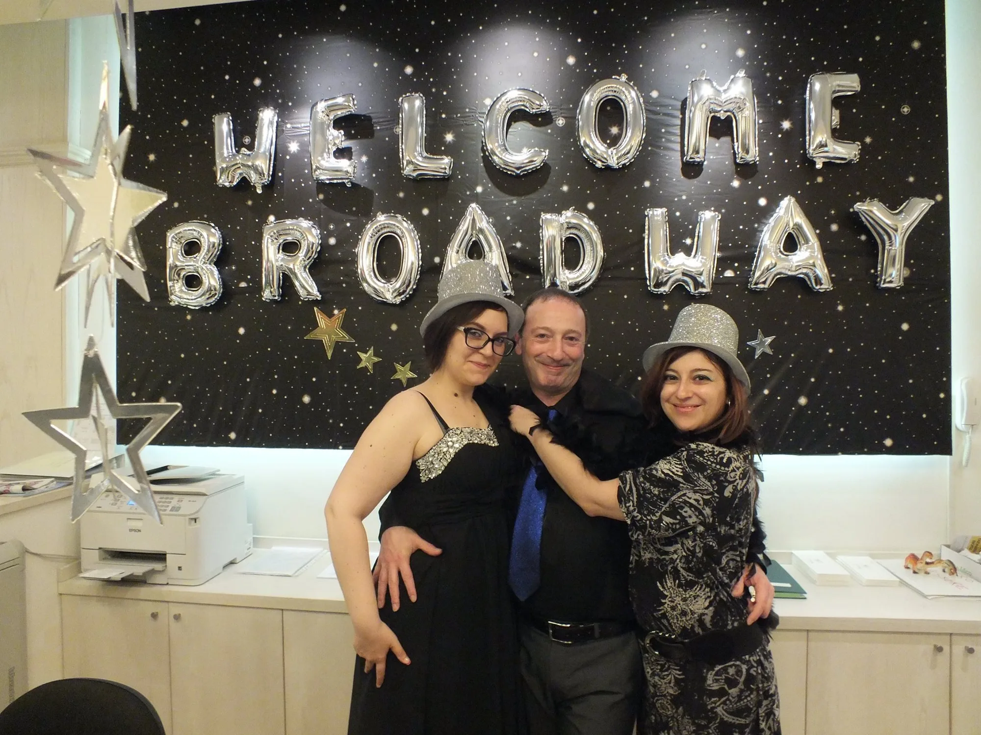 Tre persone con cappelli a cilindro argentati davanti a un fondale nero stellato con la scritta WELCOME BROADWAY in lettere di mylar argento