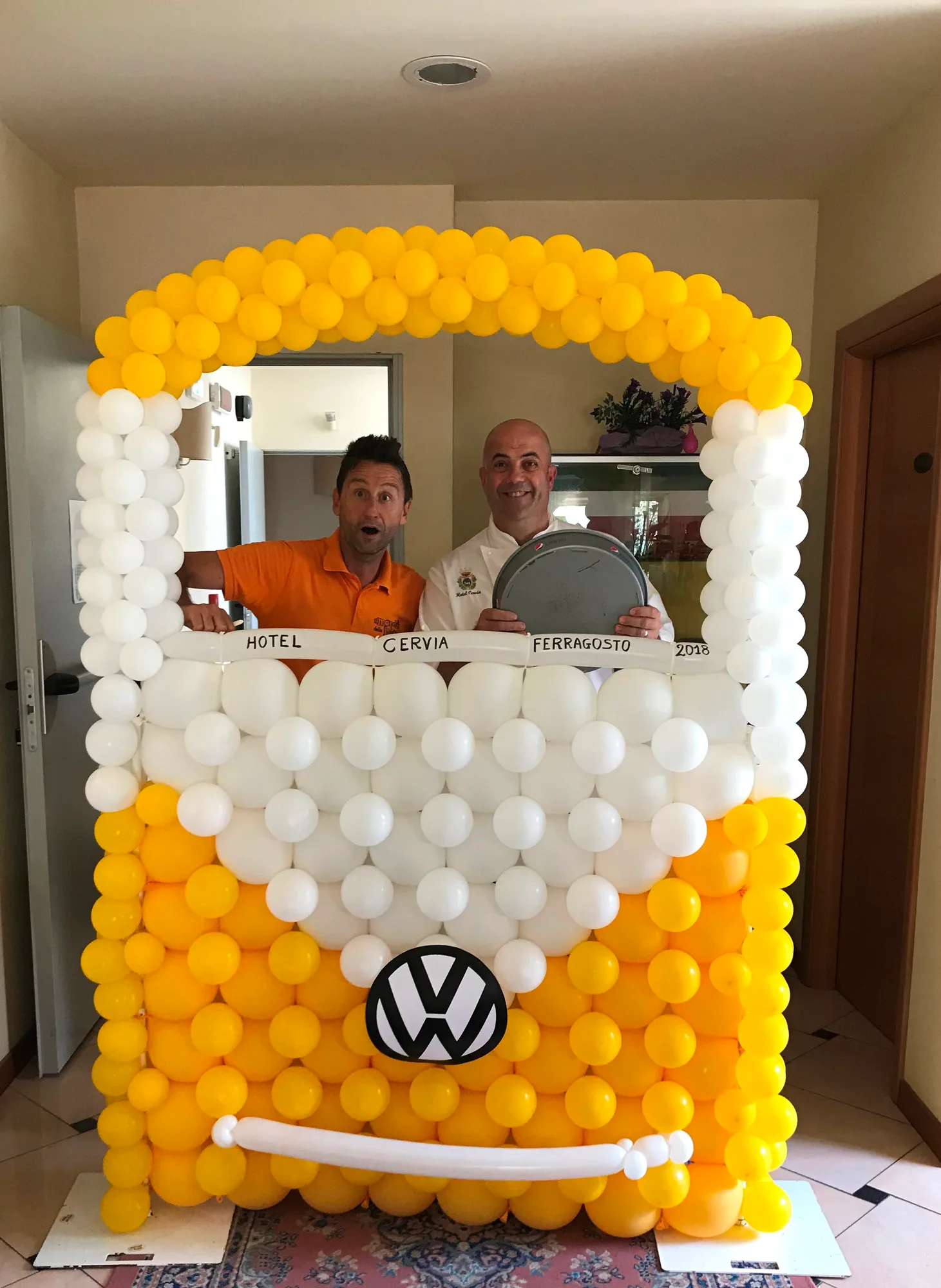 Cornice di palloncini gialli e bianchi a forma di pulmino con il logo VW e la scritta Hotel Cervia Ferragosto 2018, con due persone in posa dentro