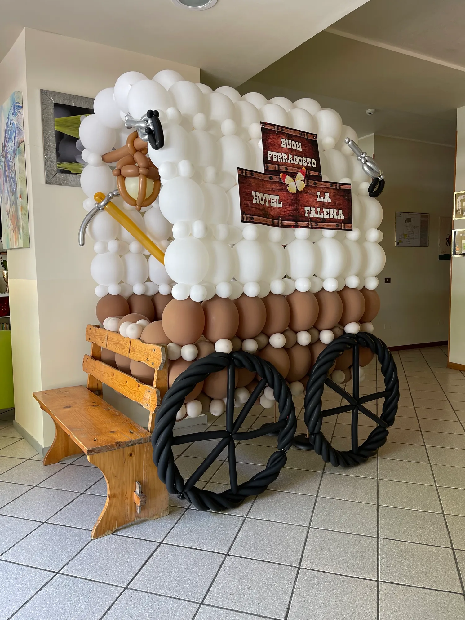 Carro del Far West di palloncini bianchi e marroni con le ruote nere, una lanterna di palloncini e il cartello Buon Ferragosto Hotel La Falena, accanto a una panca di legno