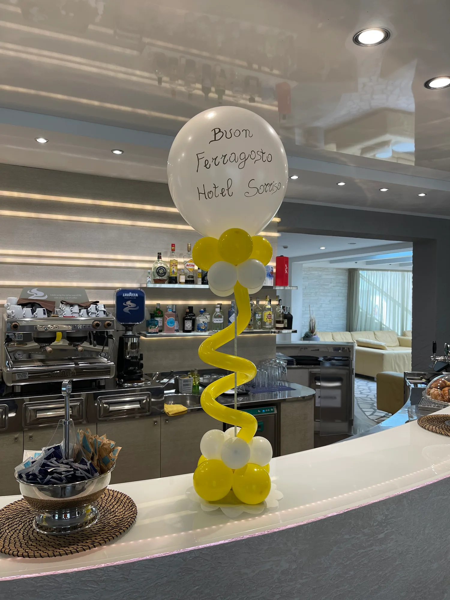Composizione di palloncini gialli e bianchi su un bancone bar, con un grande palloncino bianco scritto a mano Buon Ferragosto Hotel Sorriso