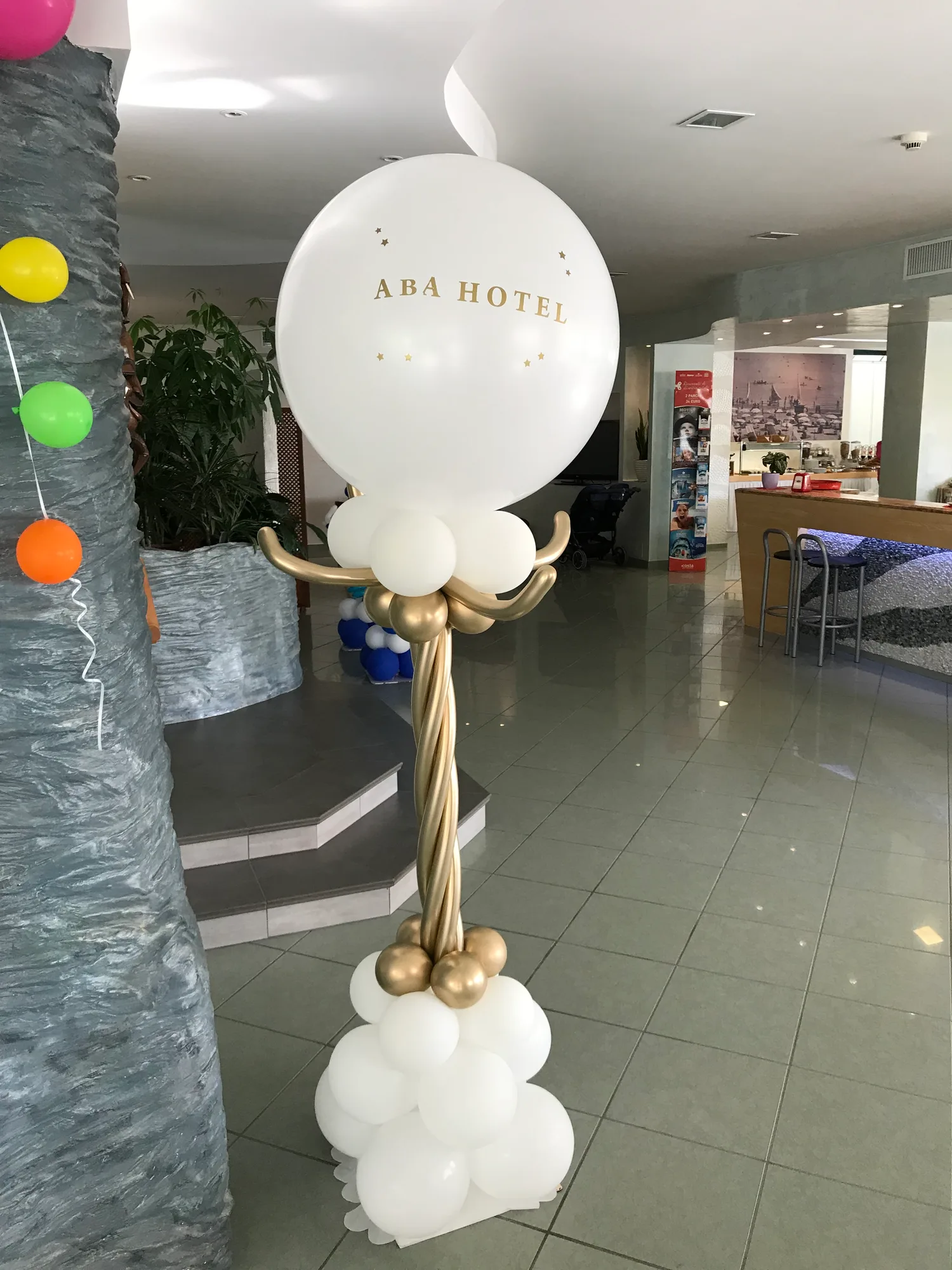 Colonna di palloncini bianchi e oro con in cima un grande palloncino bianco con la scritta ABA HOTEL, nella hall di un albergo