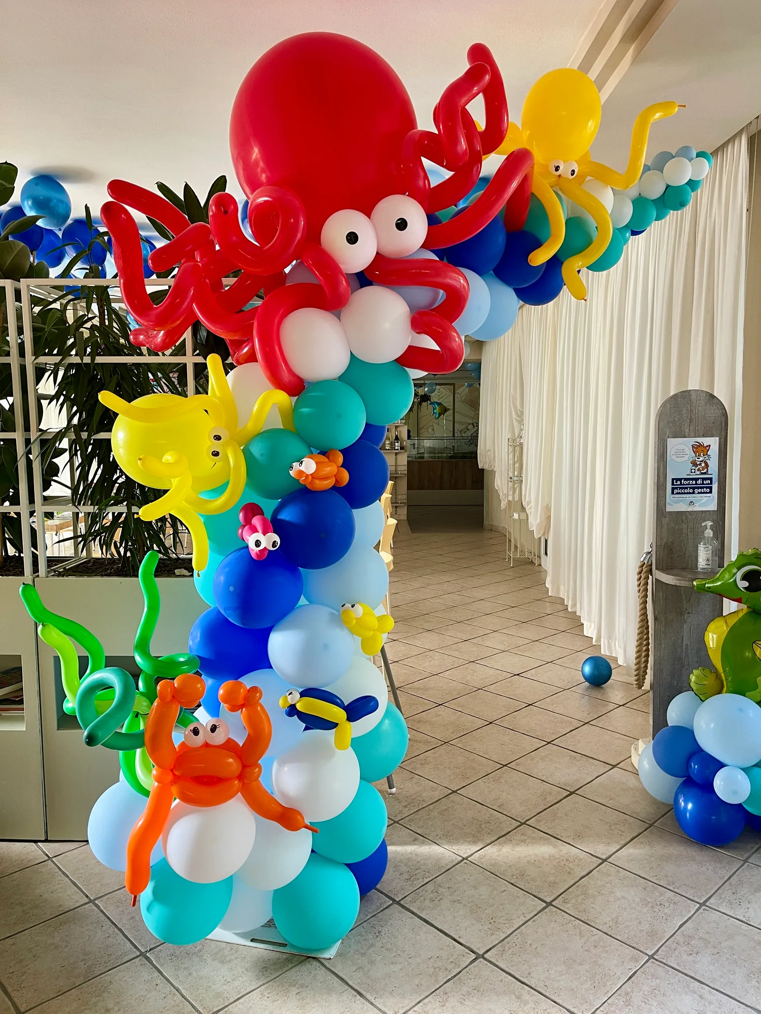 Colonna di palloncini azzurri, blu e bianchi a tema mare con un polpo rosso gigante, polpi gialli, pesciolini e un granchio, all'ingresso di una sala