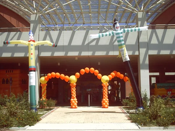 Due fly man gonfiabili, un clown e un omino a righe, ai lati dell'ingresso di un centro commerciale con arco di palloncini arancioni