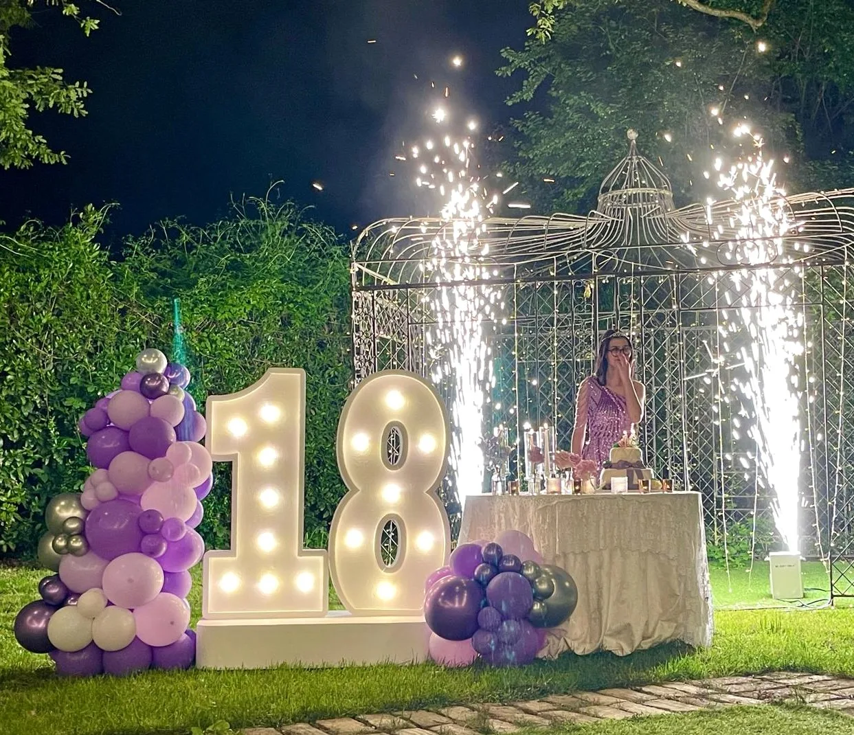 Fontane scintillanti dietro al tavolo della torta di un diciottesimo in giardino, con il numero 18 luminoso e colonne di palloncini lilla