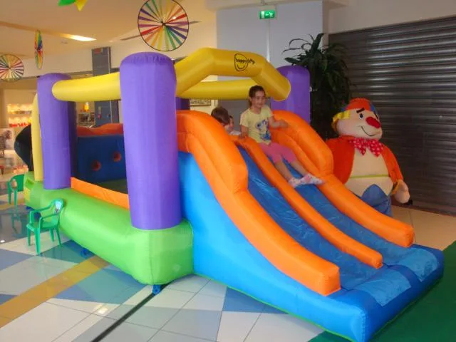 Gonfiabile Baby Scivolino montato in una galleria commerciale: due scivoli arancioni e azzurri, colonne viola, archi gialli e il grande pupazzo del clown a lato, con due bambine che scendono