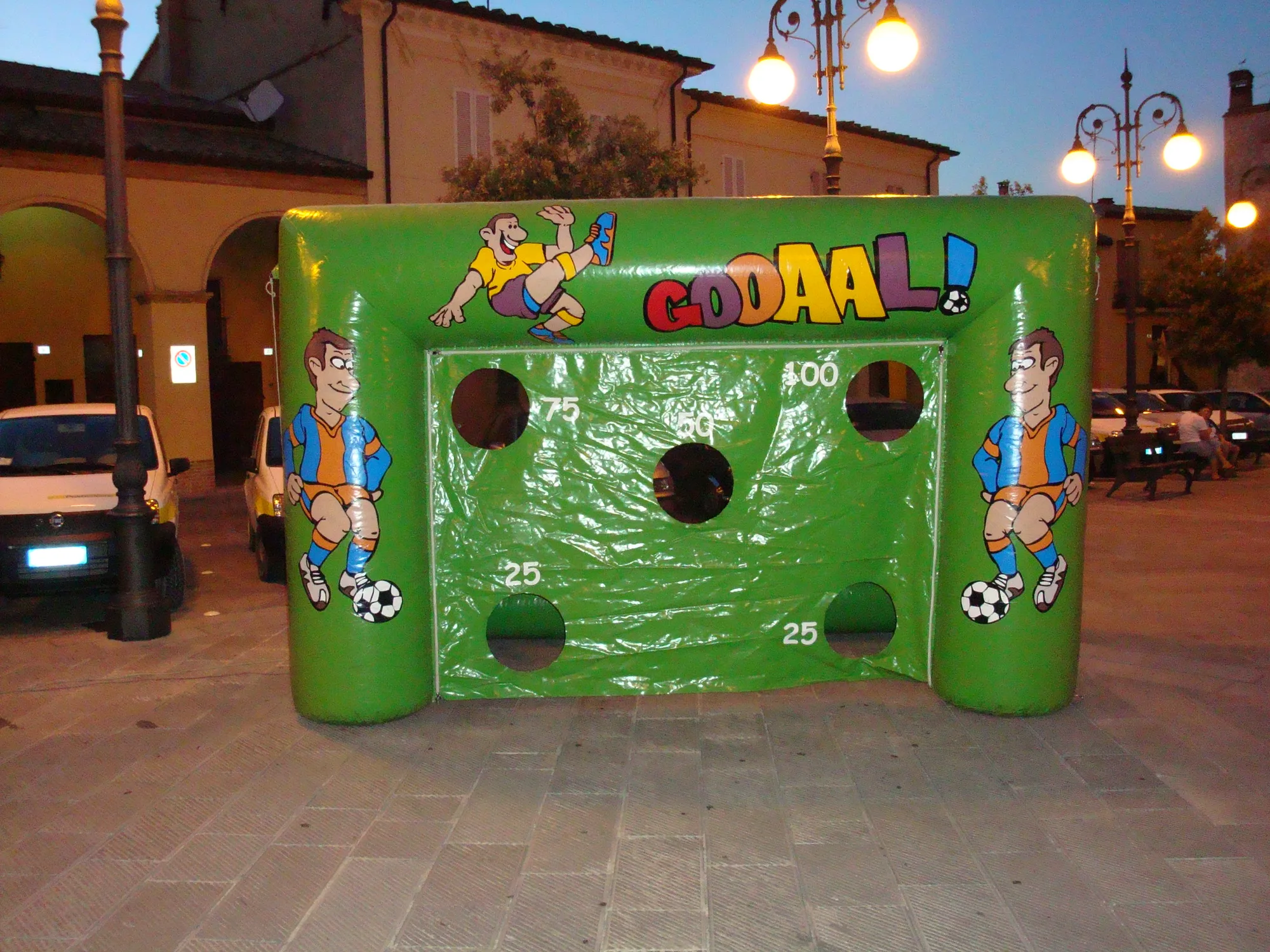 Porta da calcio gonfiabile verde Gooaal con i fori da 25, 50, 75 e 100 punti e i calciatori disegnati ai lati, montata in una piazza la sera