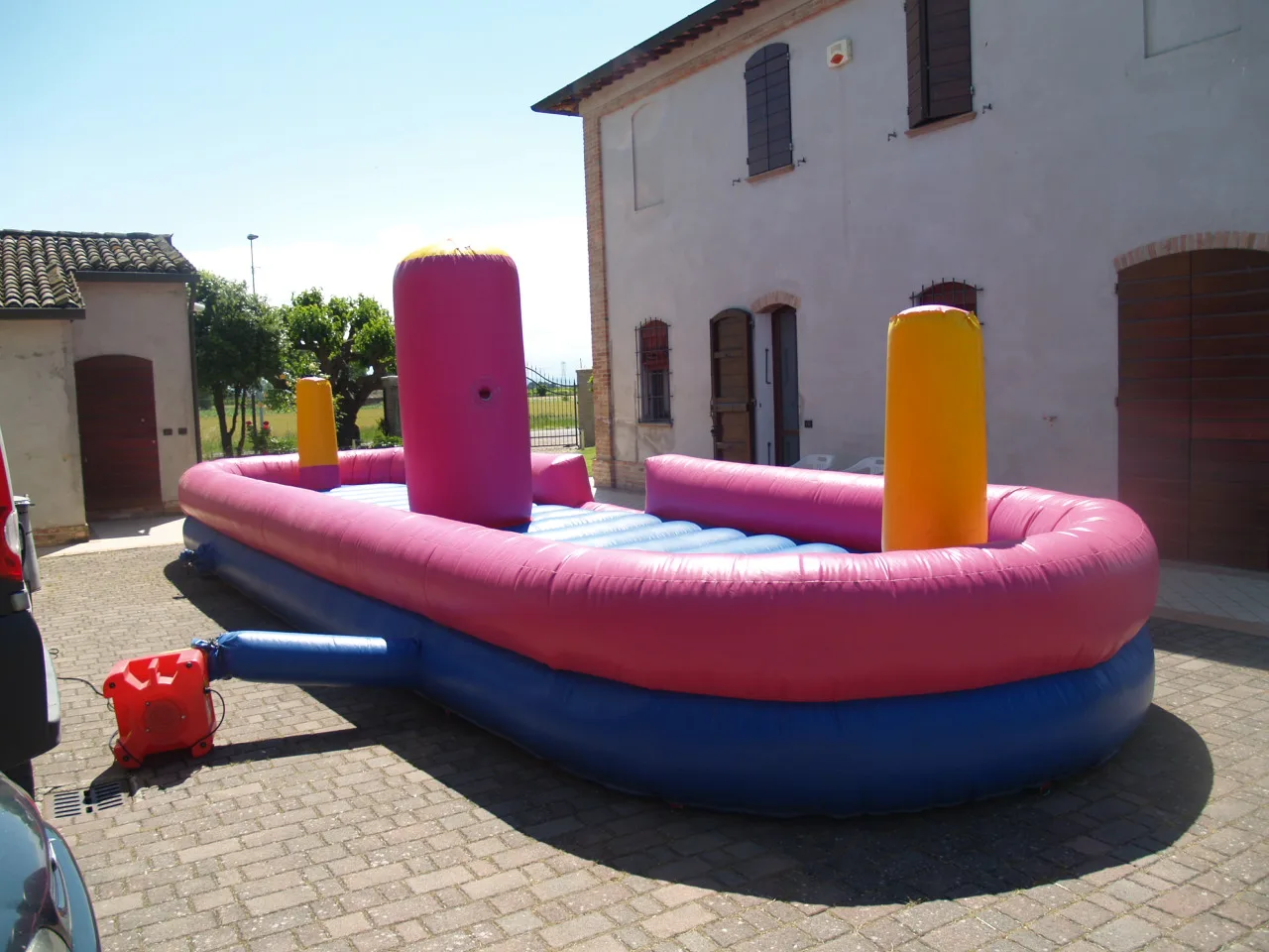 Pista gonfiabile Tira e Molla ovale, rosa e blu, con i pali morbidi gialli e rosa al centro e il gonfiatore rosso collegato, montata in un cortile