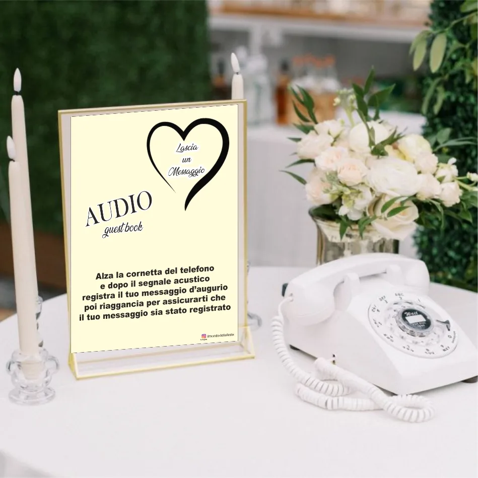 Telefono bianco vintage su un tavolo apparecchiato con tovaglia chiara, candele e un mazzo di rose bianche, accanto al cartello «Audio guest book — lascia un messaggio» in cornice dorata