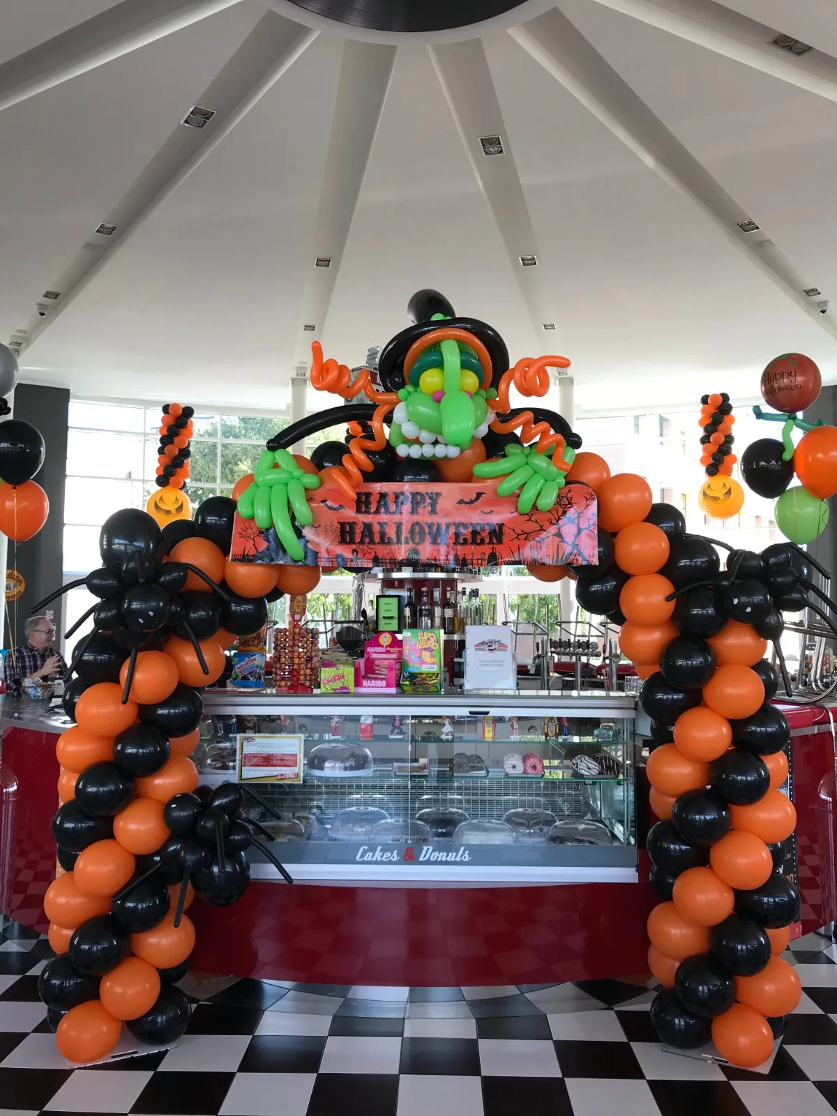 Arco di palloncini arancio e neri con ragni di palloncini sulle colonne e una strega verde in cima che regge lo striscione Happy Halloween, davanti al bancone di un locale