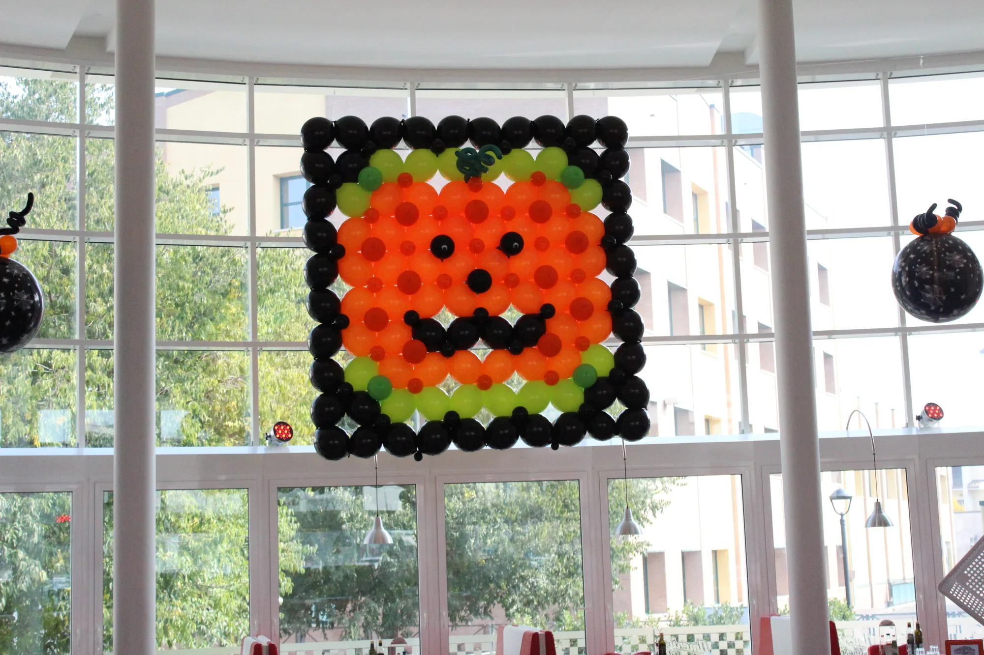 Pannello quadrato di palloncini con il bordo nero e una zucca arancione sorridente, appeso davanti alle vetrate di un locale