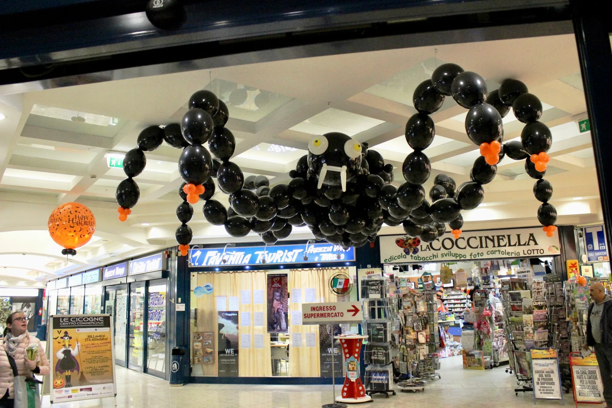Ragno gigante di palloncini neri con gli occhi gialli, i denti bianchi e le zampe con i piedi arancioni, appeso nella galleria di un centro commerciale