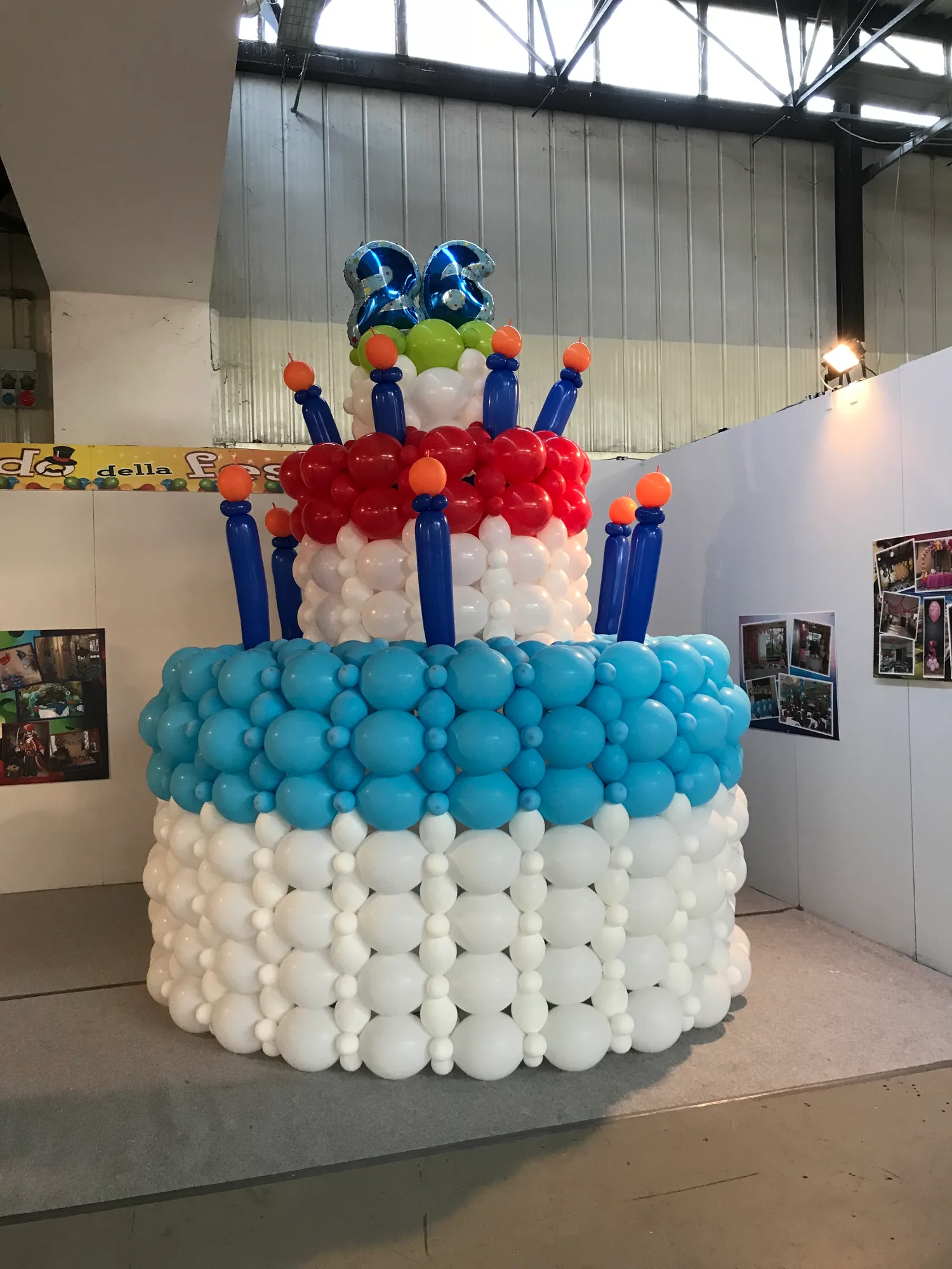 Torta gigante di palloncini bianchi con confetti colorati e candeline, appesa al soffitto della galleria di un centro commerciale con un numero 9 blu in mylar sopra