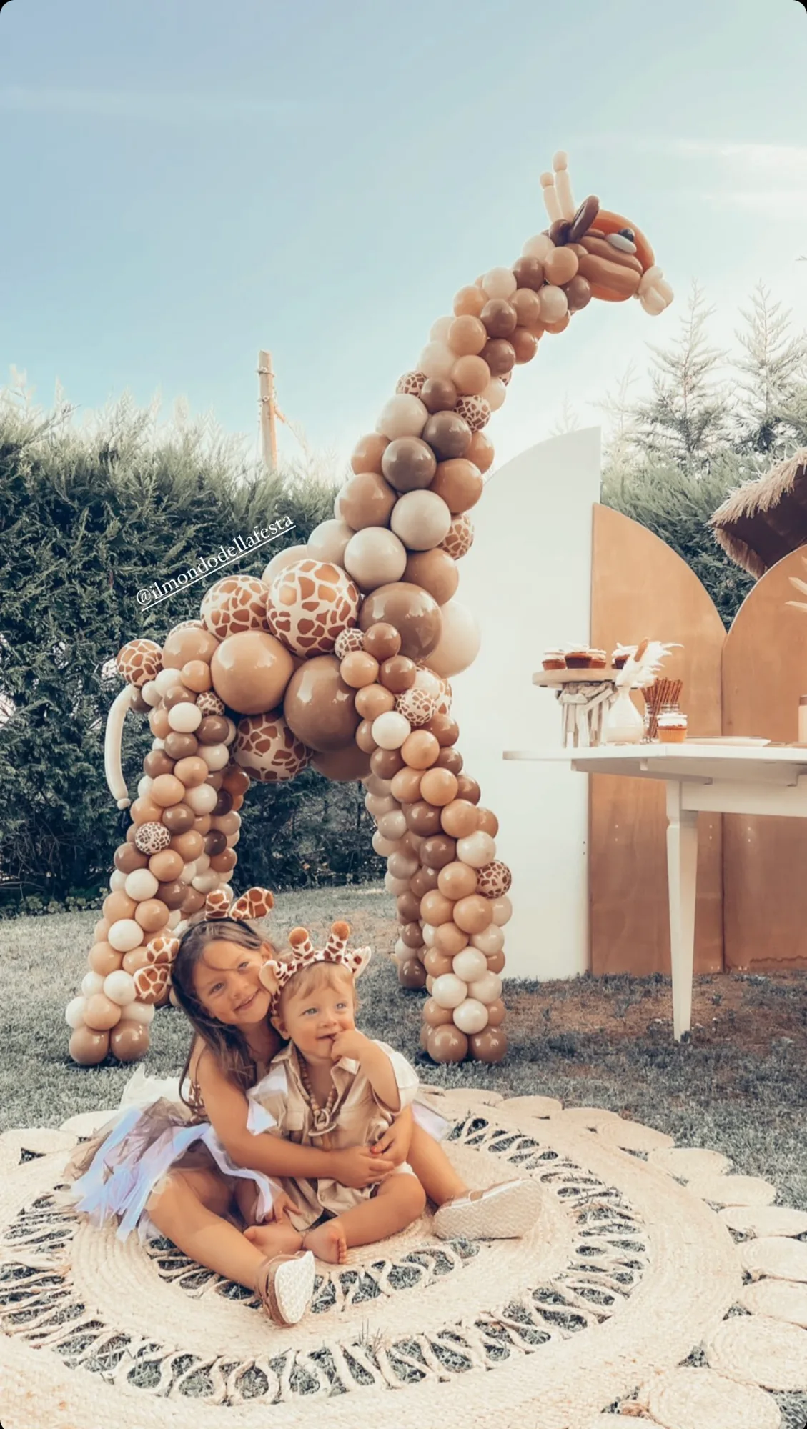 Giraffa gigante di palloncini color sabbia, marrone e maculati, alta più di due metri in un giardino allestito per una festa, con due bambini con le orecchie da giraffa seduti su un tappeto davanti