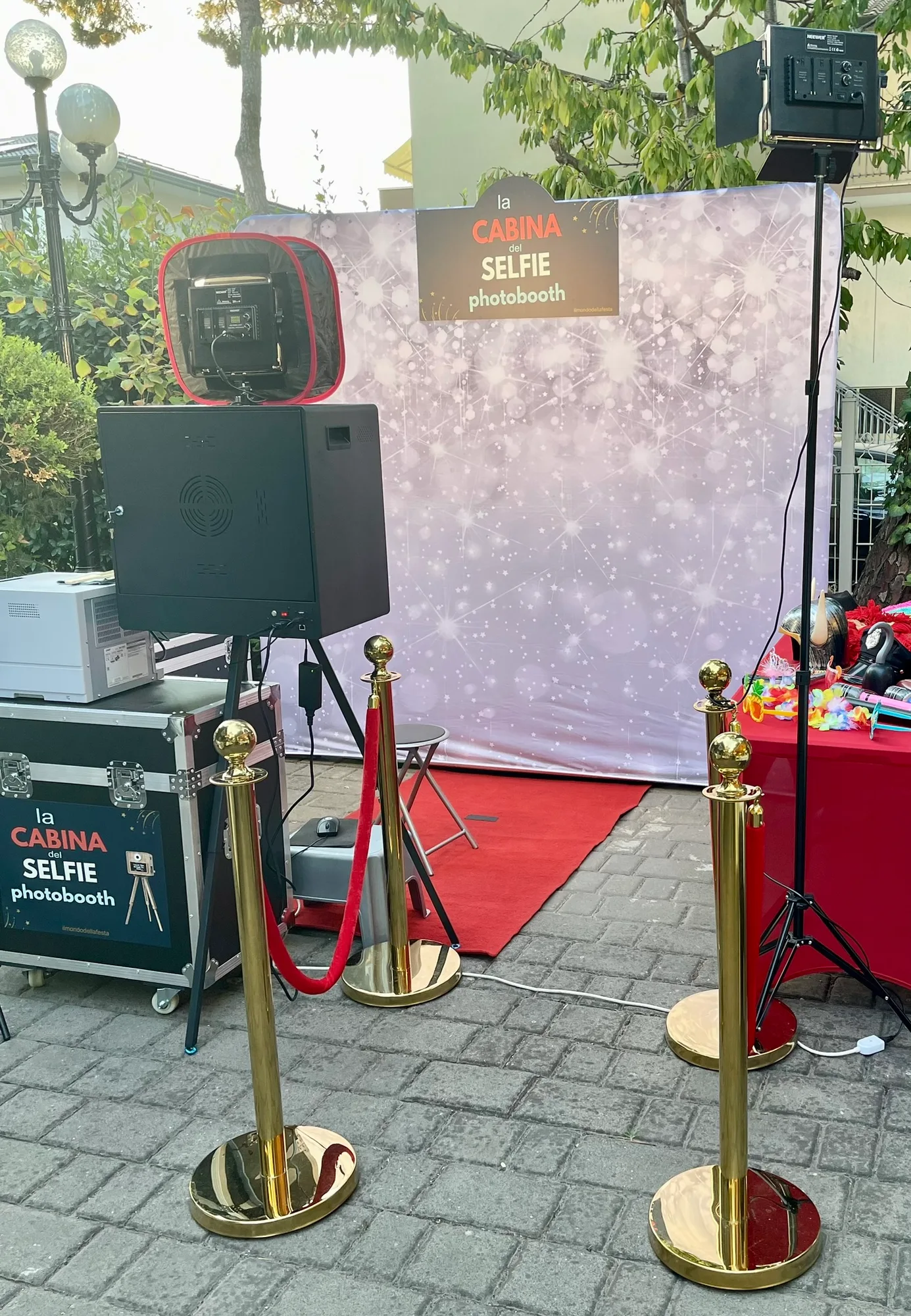 La Cabina del Selfie montata in giardino: fondale bianco scintillante con l'insegna, tappeto rosso, colonnine dorate con cordone rosso e la cabina fotografica nera su treppiede con faretto