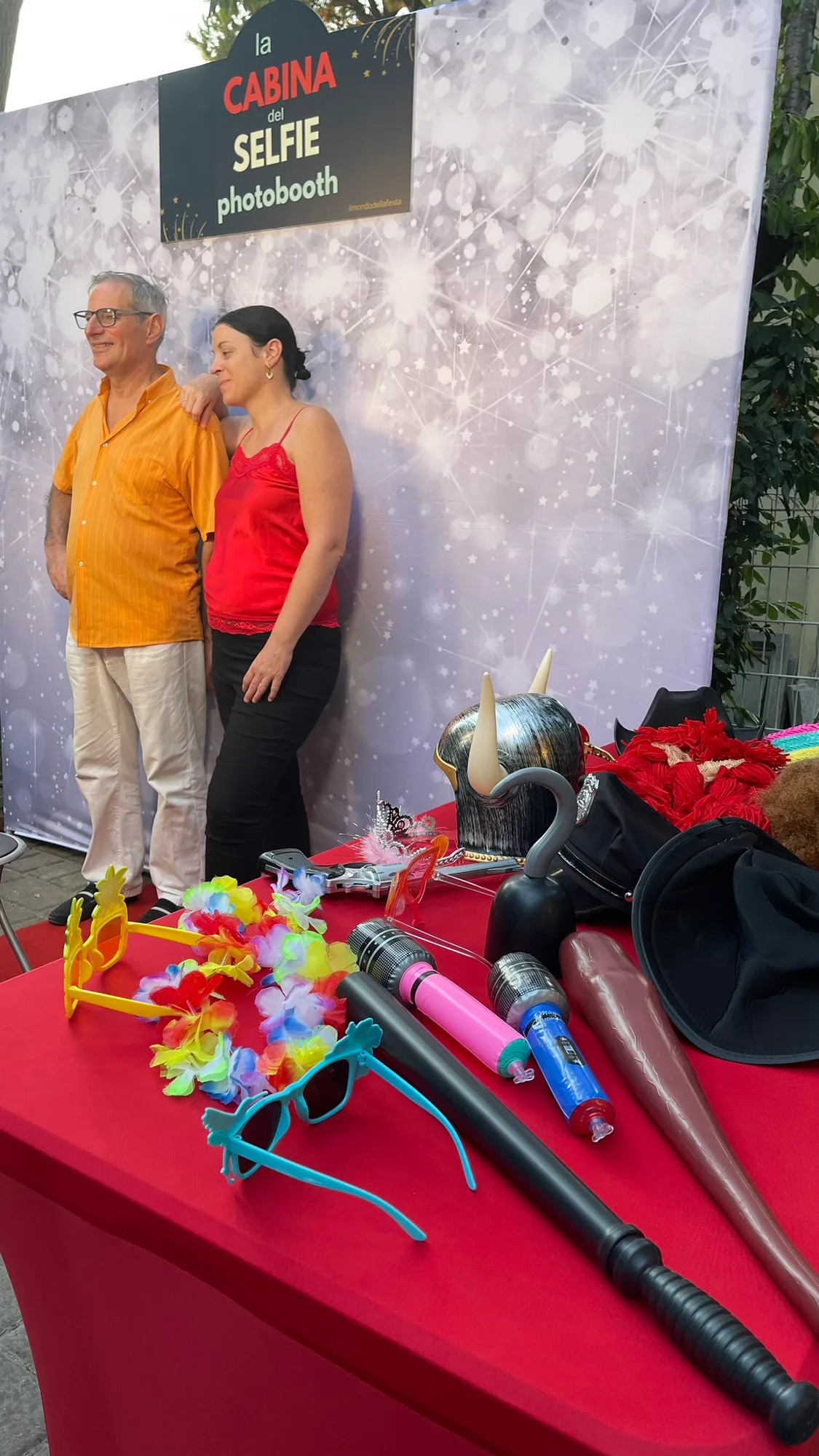 Coppia di invitati in posa davanti al fondale della Cabina del Selfie, con il tavolo rosso degli accessori in primo piano