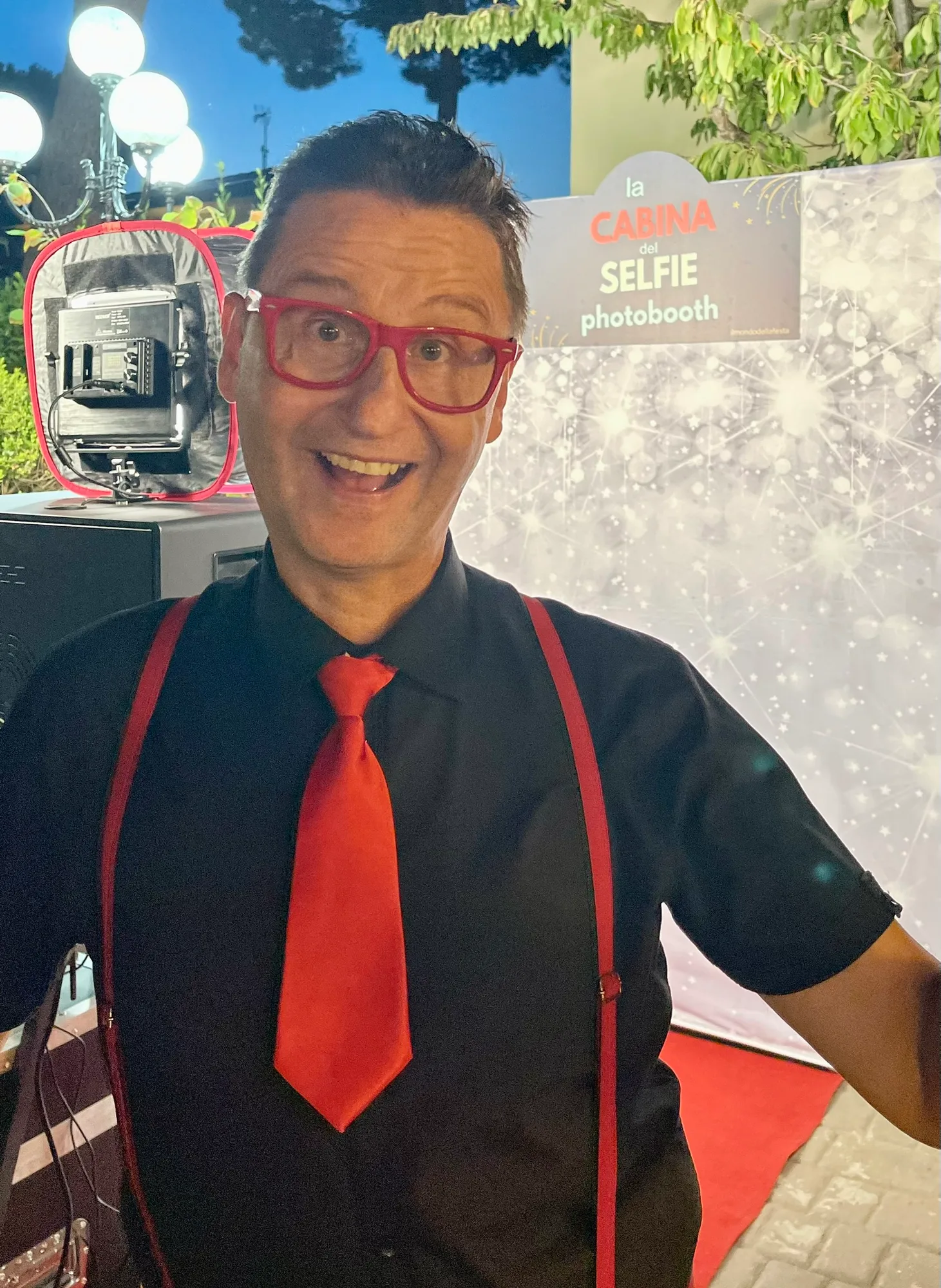 L'addetto della Cabina del Selfie in camicia nera, bretelle e cravatta rossa davanti al fondale scintillante, con la cabina e il faretto accesi alle spalle