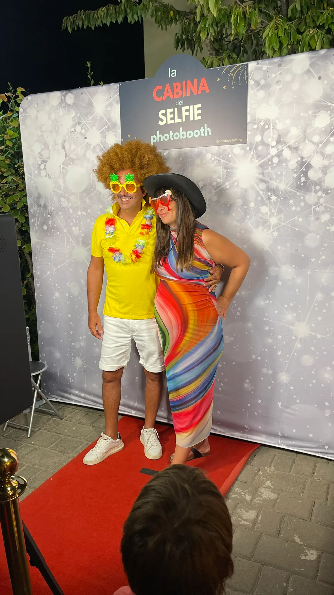 Coppia di invitati in posa con parrucca riccia, occhiali a forma di ananas, collana hawaiana e cappello nero davanti al fondale del photobooth