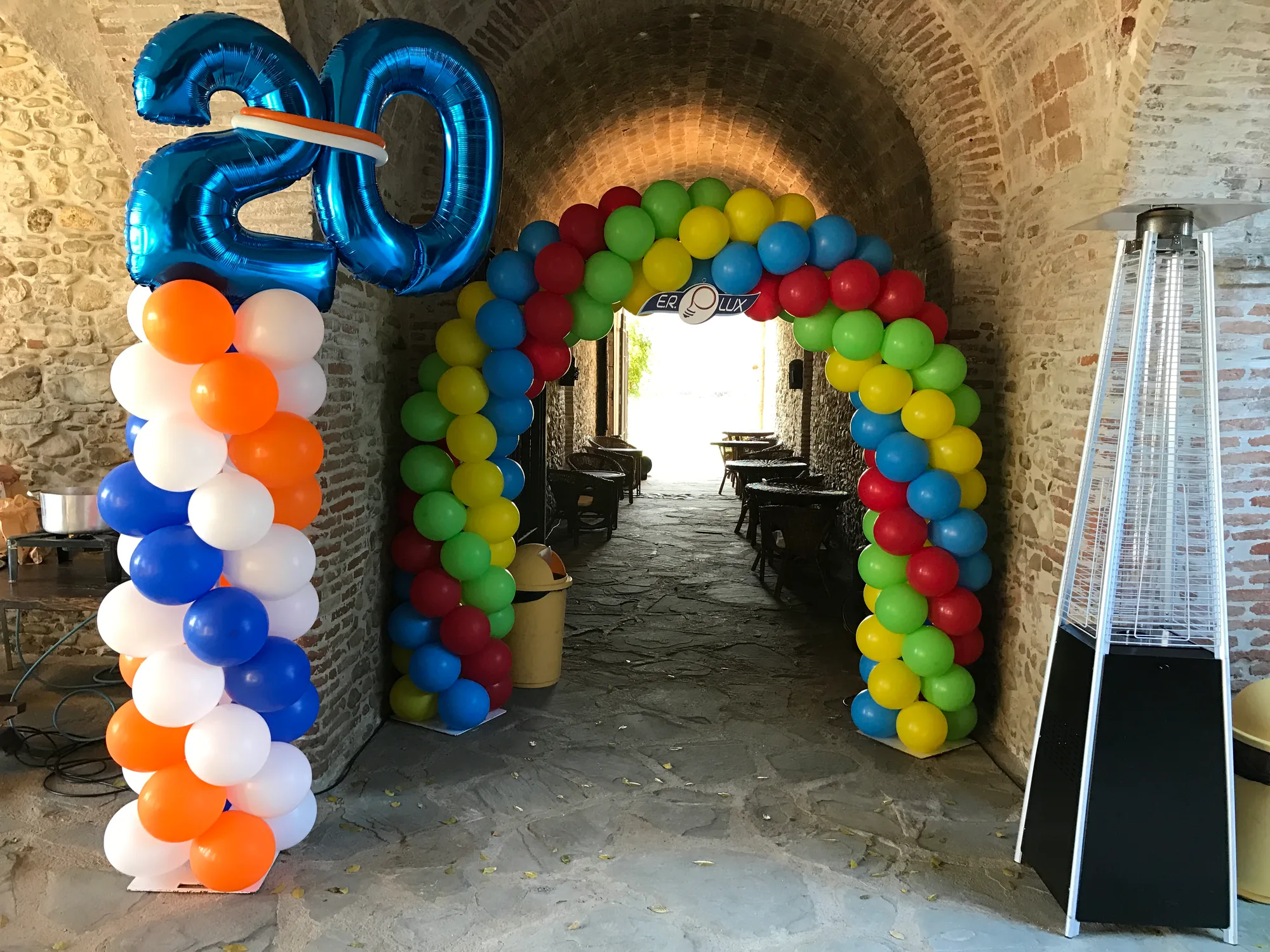 Arco di palloncini multicolore sotto una volta in mattoni con il logo dell'azienda appeso al centro, numero 20 in mylar blu e colonna arancione, bianca e blu per la festa dei vent'anni di attività