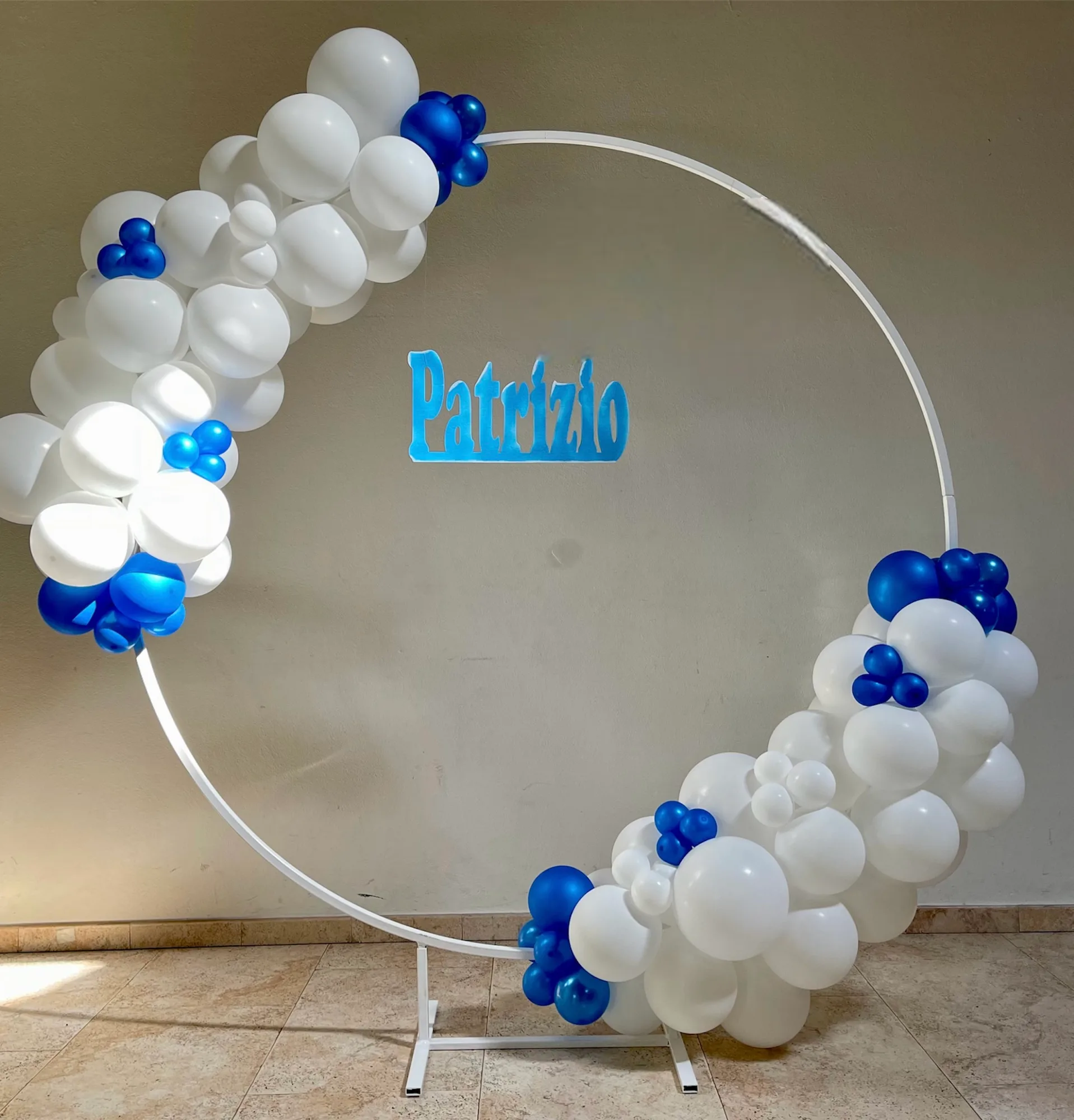 Cerchio di palloncini bianchi e blu montato su struttura tonda, con la sagoma del nome Patrizio applicata alla parete