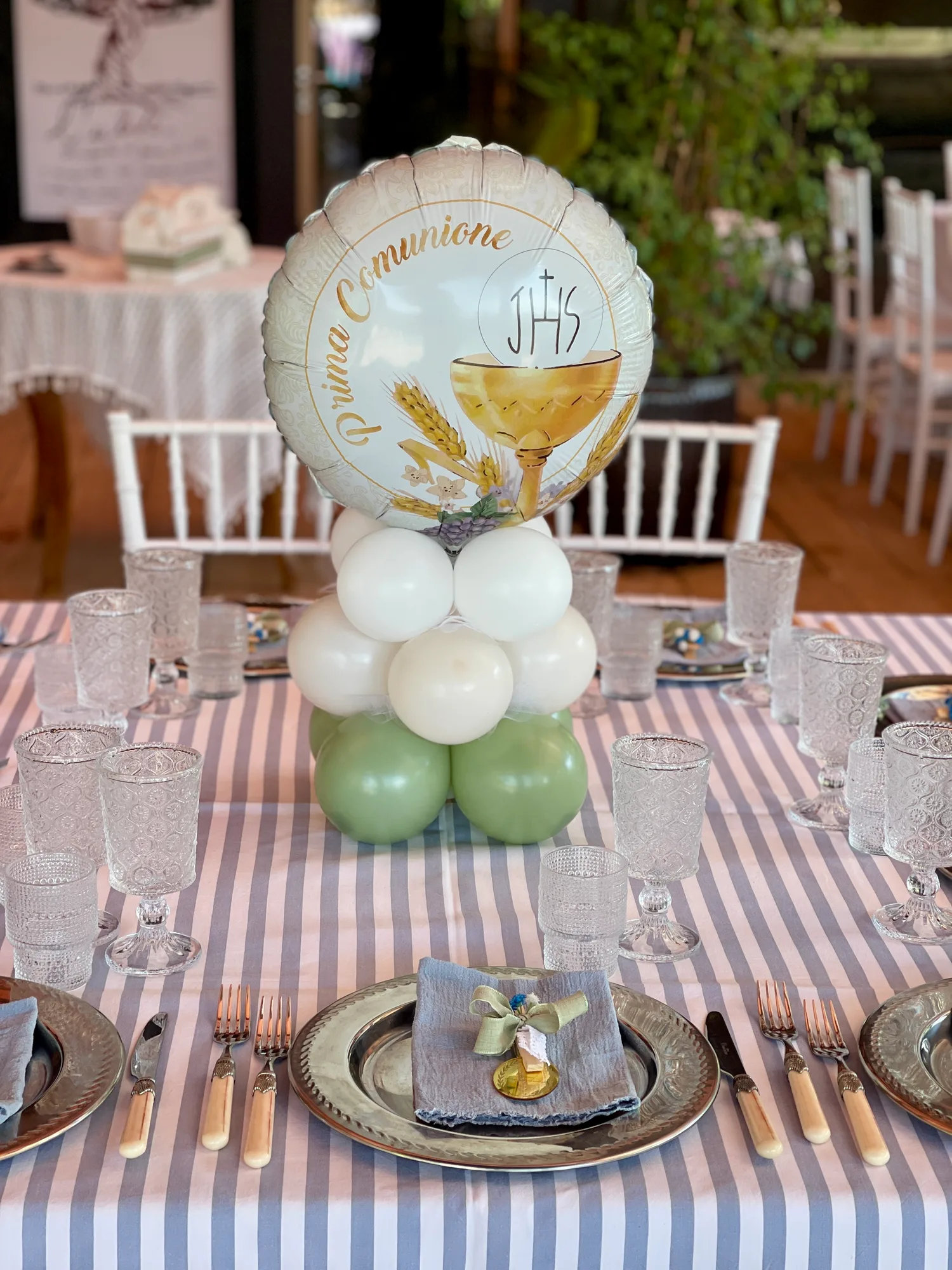 Centrotavola con palloncino Prima Comunione decorato con calice e spighe, su una base di palloncini verde salvia e panna, sopra la tavola apparecchiata