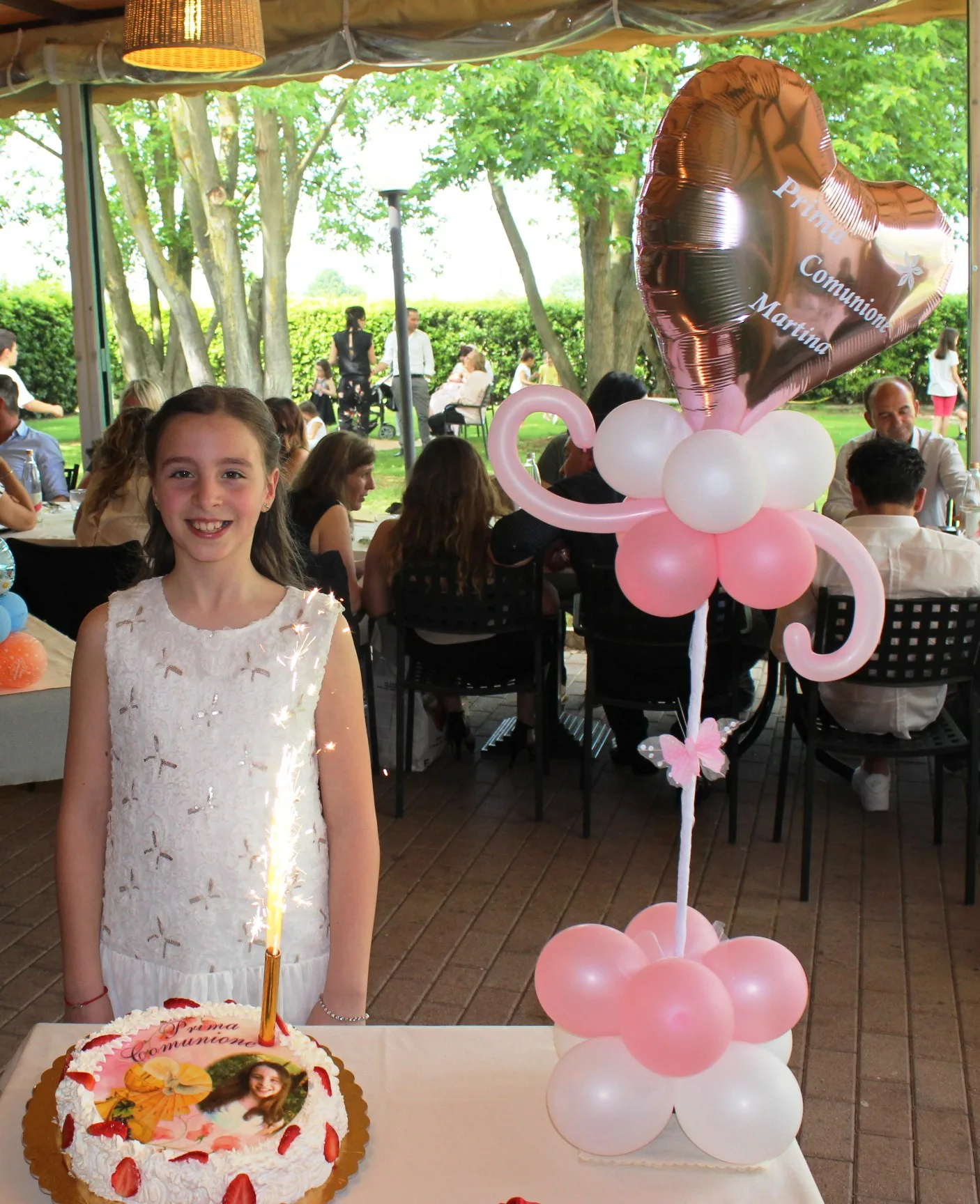 Bambina accanto alla torta della Prima Comunione e a una composizione a fiore di palloncini rosa e bianchi con cuore in mylar rose gold personalizzato Prima Comunione Martina