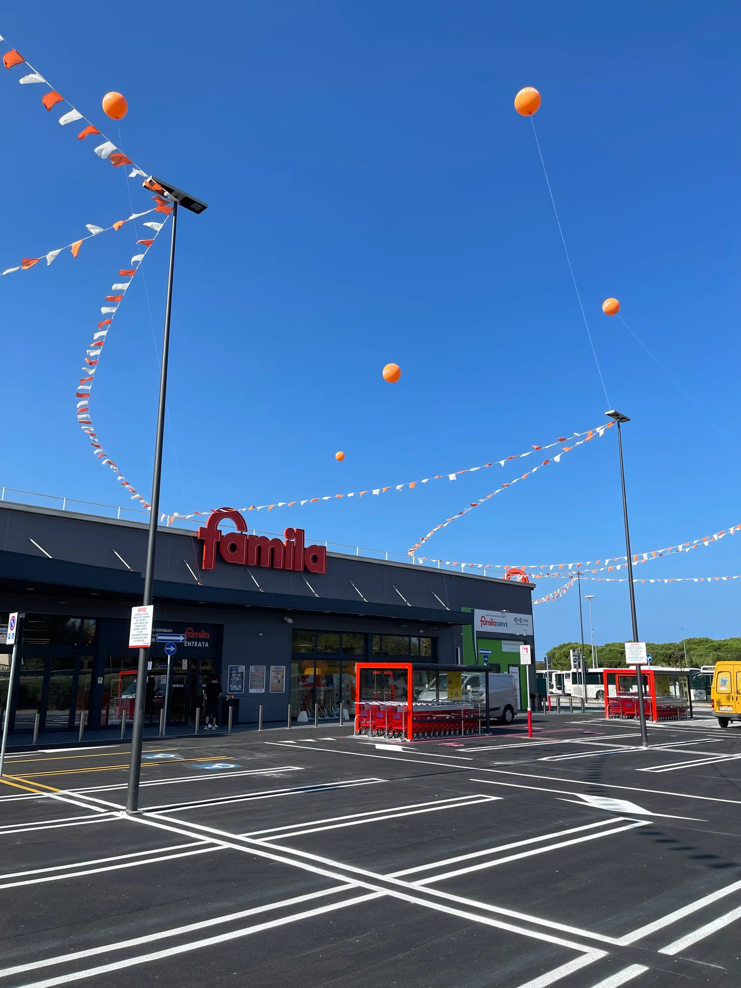 Bandierine arancioni e bianche tese sopra il parcheggio di un supermercato e palloncini giganti a elio ancorati ai lampioni contro il cielo azzurro