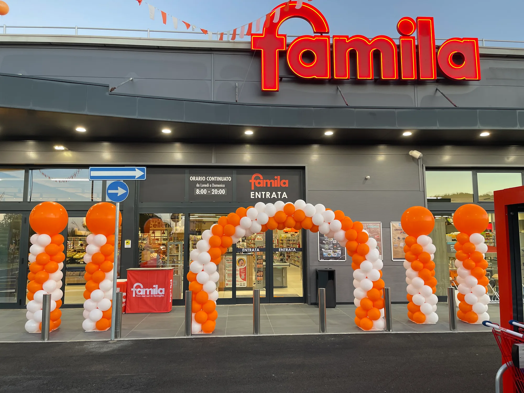 Arco di palloncini arancioni e bianchi sopra le porte di ingresso di un supermercato appena aperto, con quattro colonne sormontate da sfere giganti e bandierine sul tetto