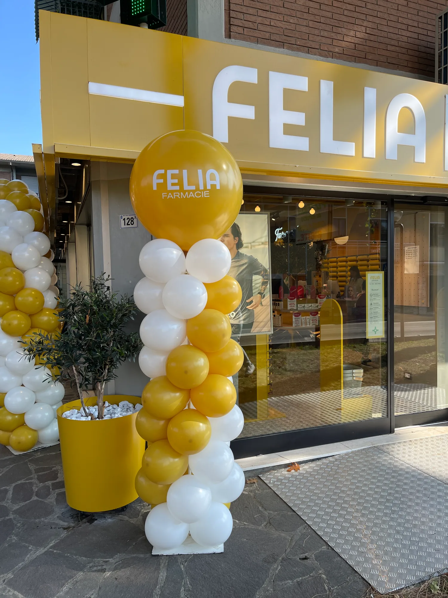 Colonne di palloncini gialli e bianchi davanti alla vetrina di una farmacia appena aperta, con sfera gialla gigante stampata con il logo dell'attività
