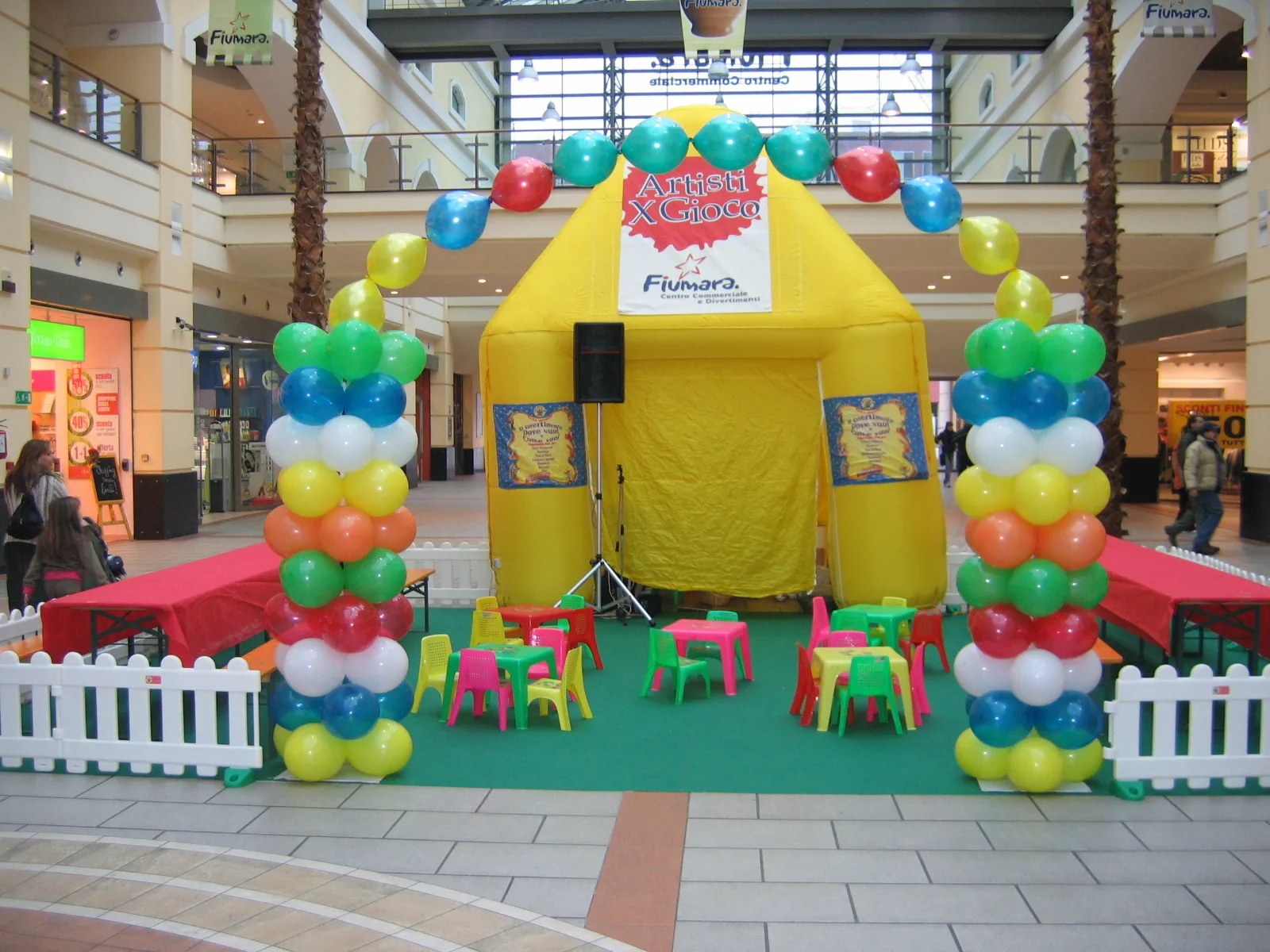 Baby parking in una galleria commerciale: gazebo gonfiabile giallo, recinzione bianca, tappeto verde con tavolini e seggioline colorate e due colonne di palloncini all'ingresso